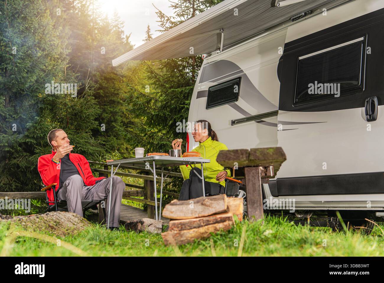 Zwei Personen sitzen an einem Tisch bei einem Wohnmobil und teilen sich bei Tageslicht in einem Waldgebiet Speisen und Getränke. Stockfoto