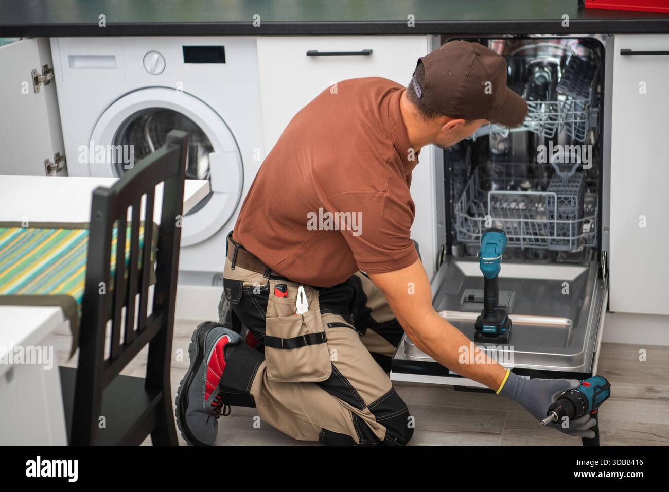 Ein Mann arbeitet daran, einen Geschirrspüler zu reparieren, während er auf dem Küchenboden kniet. Werkzeuge befinden sich in der Nähe auf dem Tresen. Stockfoto