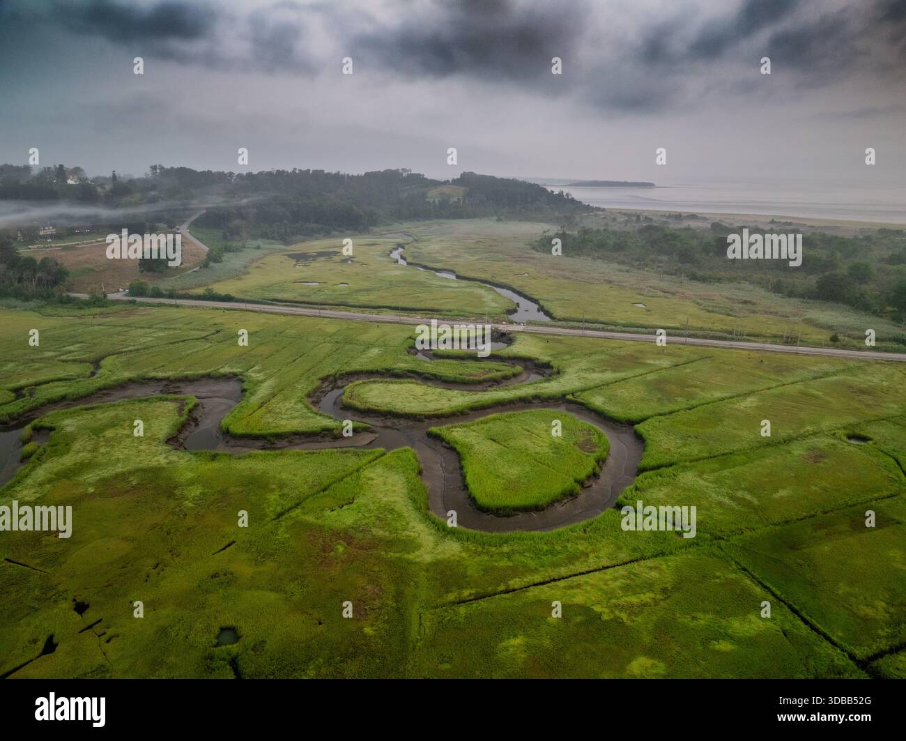 Aus der Vogelperspektive schlängelnde Buchten schlängeln sich durch das pulsierende grüne Sumpfland unter einem glühenden Himmel und kontrastieren den ruhigen Horizont des Meeres, Gloucester, Mas Stockfoto