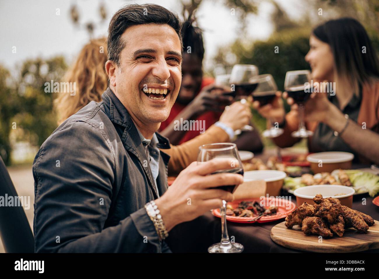 Ehrliches Porträt eines lächelnden Mannes, der Wein bei einem Abendessen im Freien mit Freunden genießt. Warme Atmosphäre, echte Emotionen, authentischer Lebensstil und fröhlich Stockfoto