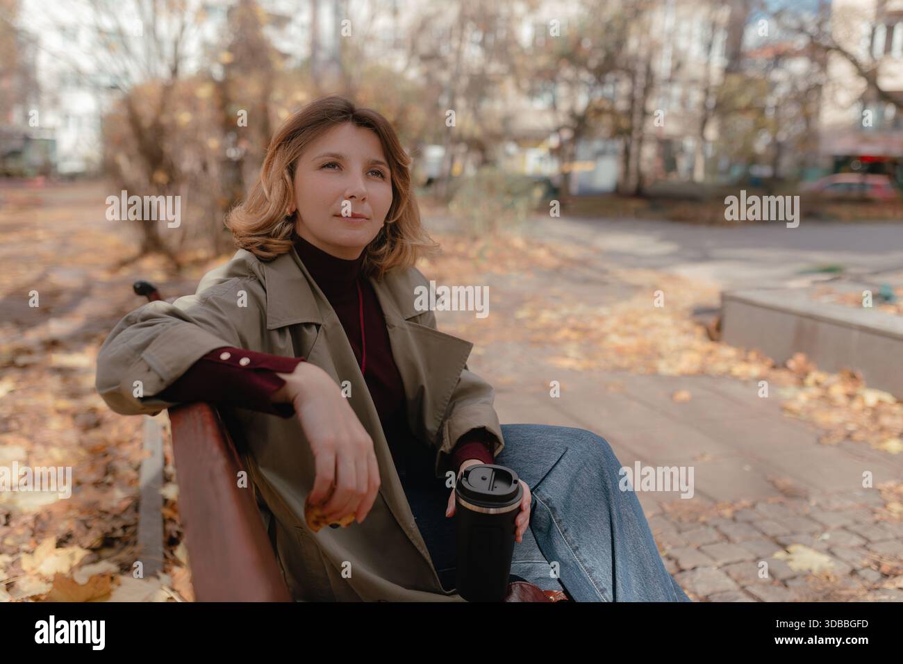 Frau sitzende Parkbank mit Reisebecher, herbstliche Blätter bedecken Boden, weiches warmes Mittagslicht. Stockfoto