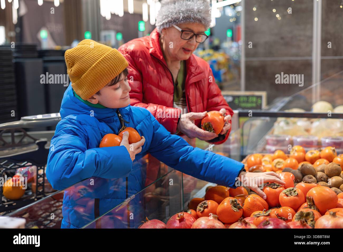 Großmutter und Enkel legen sich auf gesunde Entscheidungen, während sie in einem modernen Lebensmittelgeschäft frische Persimmons kaufen. Generationsübergreifender Lebensstil. Stockfoto