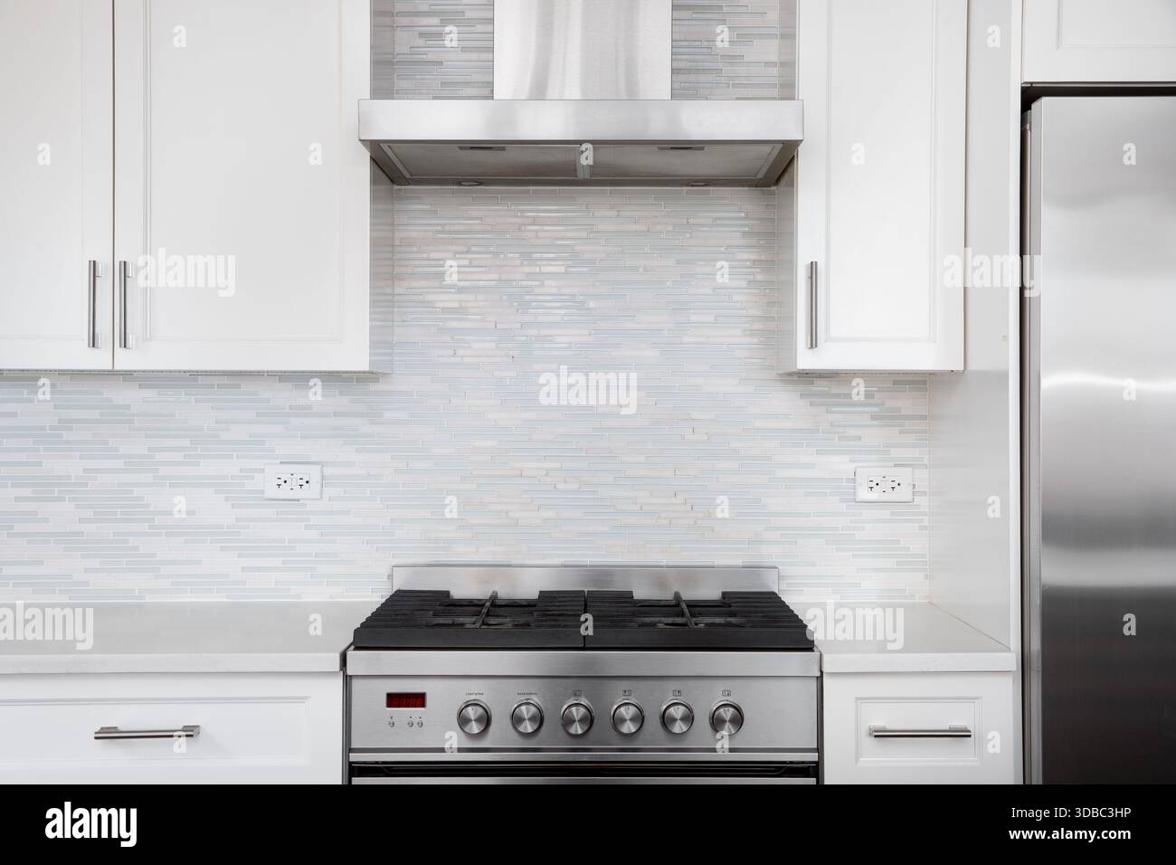 Die moderne weiße Küche verfügt über einen Gasherd aus Edelstahl gegen einen hellblauen und weißen Mosaikfliesen. Stockfoto