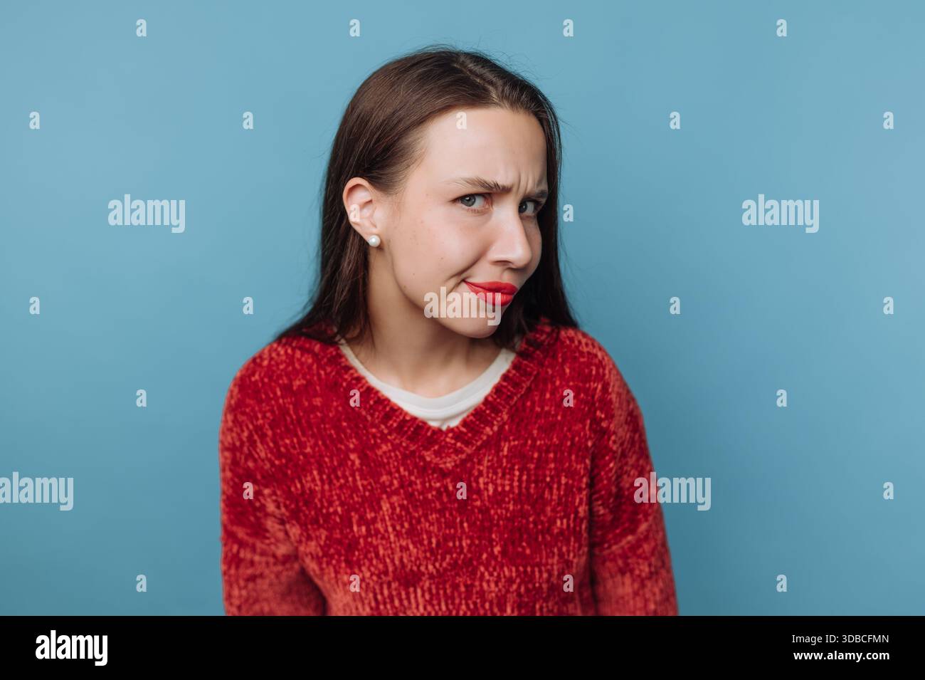 Eine junge Frau steht vor einer blauen Wand und macht einen lustigen Ausdruck. Sie trägt einen roten Pullover und hat einen verspielten Blick im Gesicht. Stockfoto