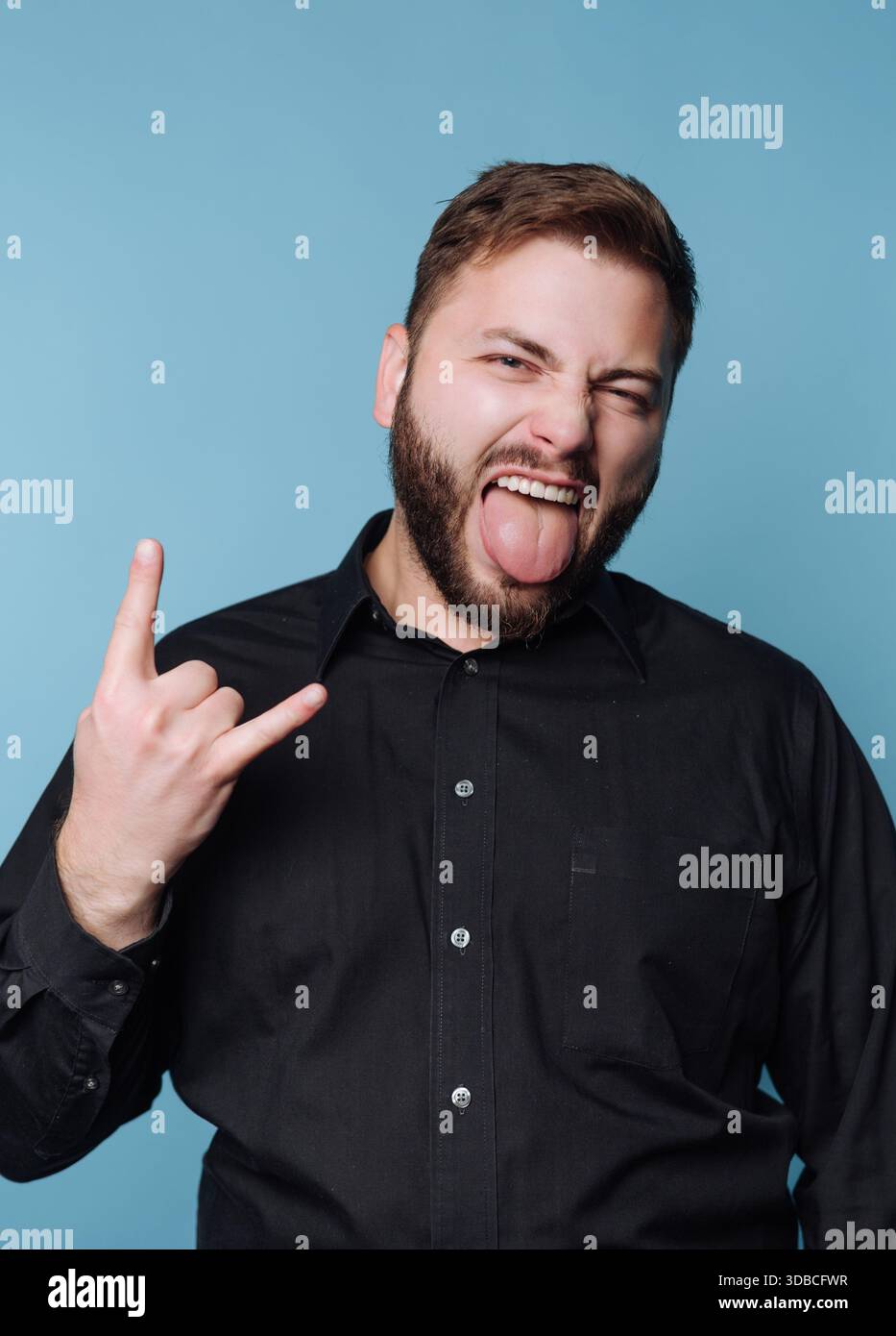 Ein Mann steht vor blauem Hintergrund. Er macht eine für Rockmusik typische Handgeste, während er seine Zunge rausstreckt. Sein Ausdruck zeigt Enthusiasmus Stockfoto