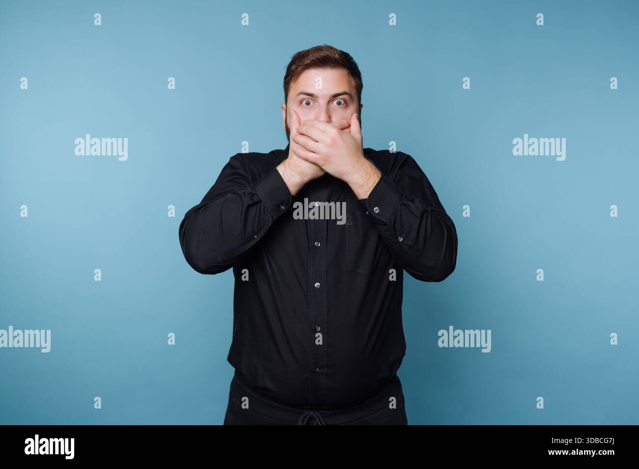 Ein Mann steht mit breiten Augen und bedeckt seinen Mund mit beiden Händen. Er scheint schockiert oder überrascht zu sein. Der Hintergrund ist einfach blau, was einen klaren Fo schafft Stockfoto