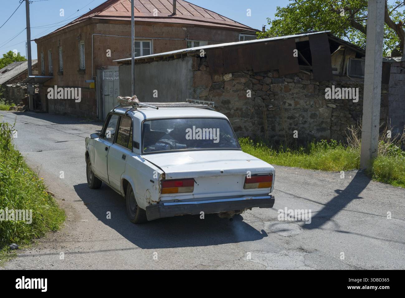 Eine Rückansicht eines alten Autos auf einer Landstraße umgeben von alten Gebäuden, Lada 2107, auch WAS-2107, VAZ-2107, Lada Nova oder Lada Riva fährt ohne LIC Stockfoto