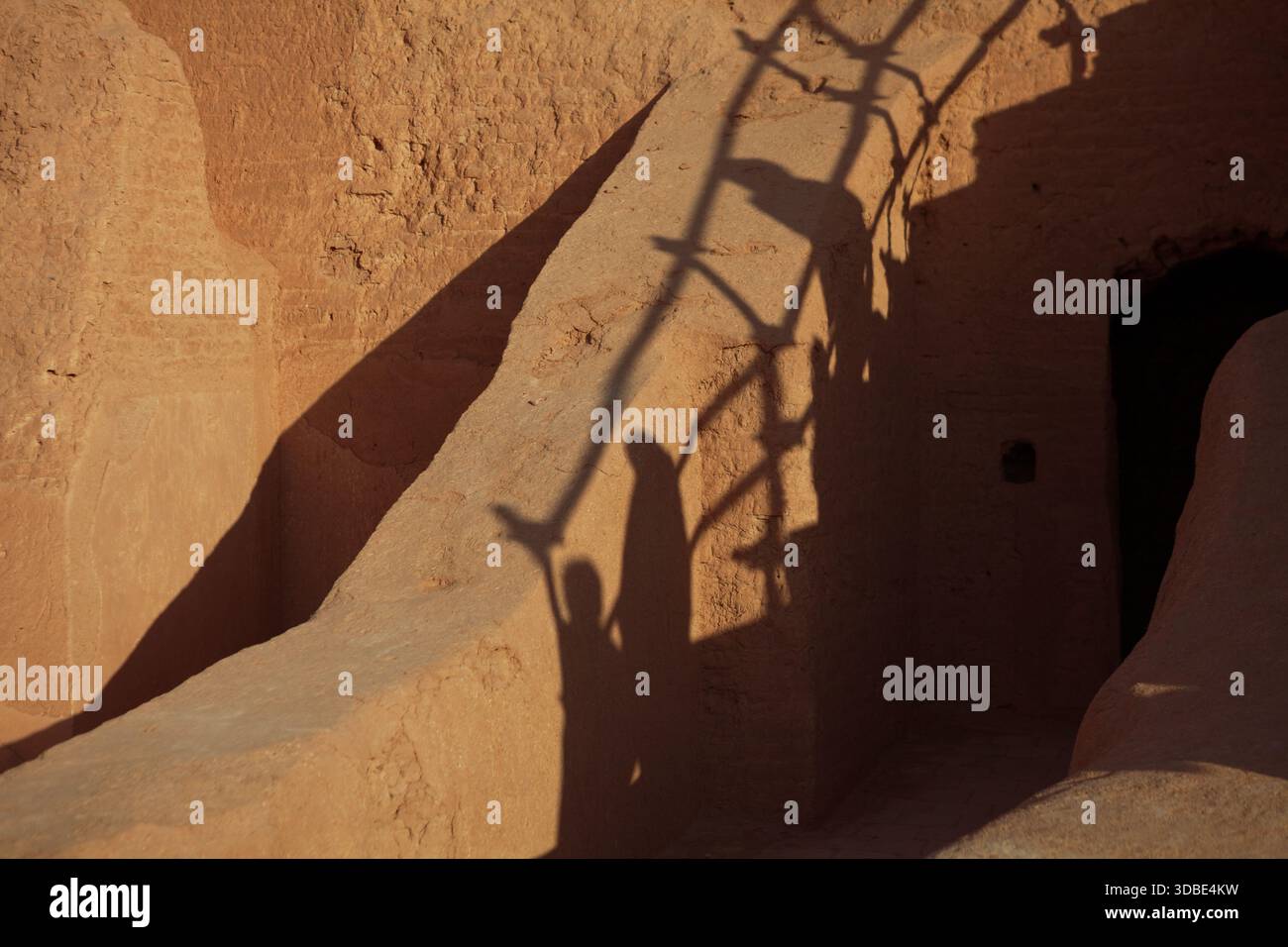 Schatten von drei Figuren und eine Leiter fallen über die sonnendurchfluteten lehmwände einer alten persischen Ruine. Warme Töne und dramatisches Licht sorgen für ein zeitloses,... Stockfoto