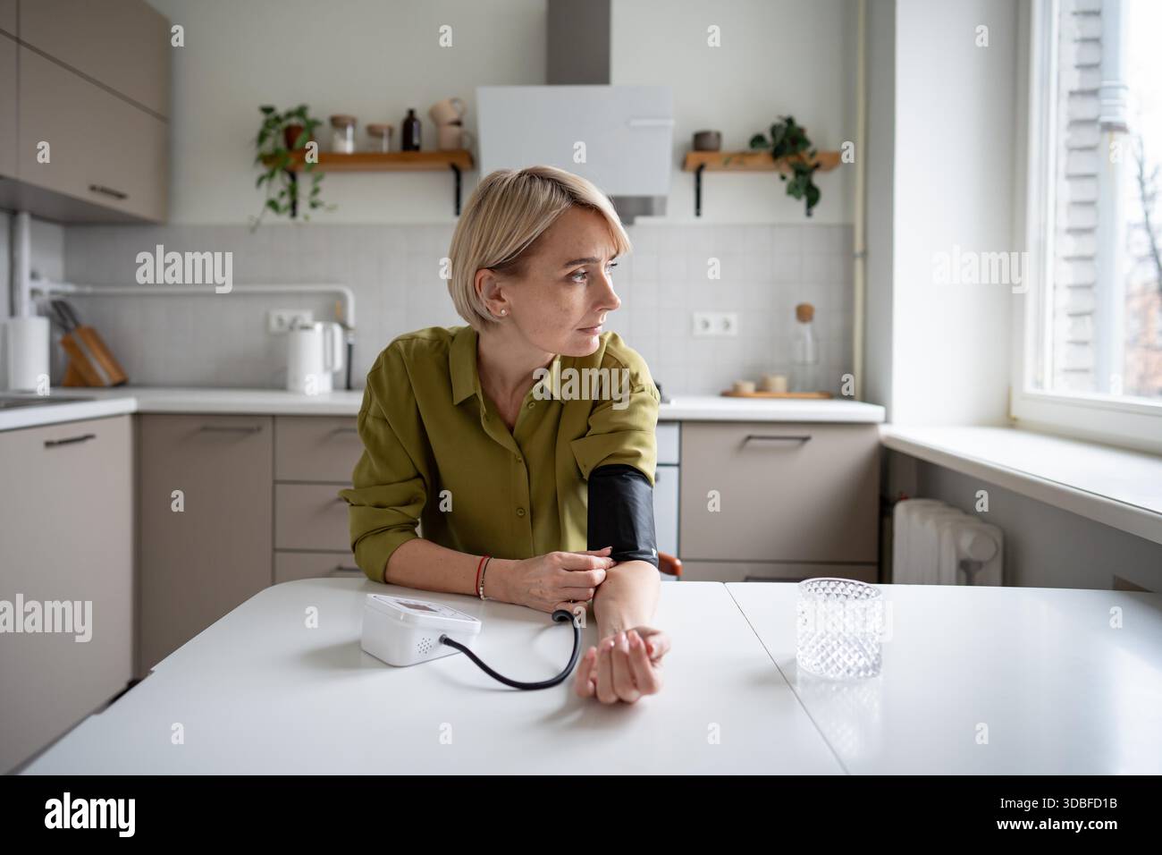 Müde Frau mittleren Alters mit erschöpfter Expression, Blutdruckmessung mit Tonometergerät, die in der Küche am Tisch sitzt. Frauen leiden unter Müdigkeit und Energiemangel, um den Druckpegel zu überprüfen. || Modell freigegeben Stockfoto