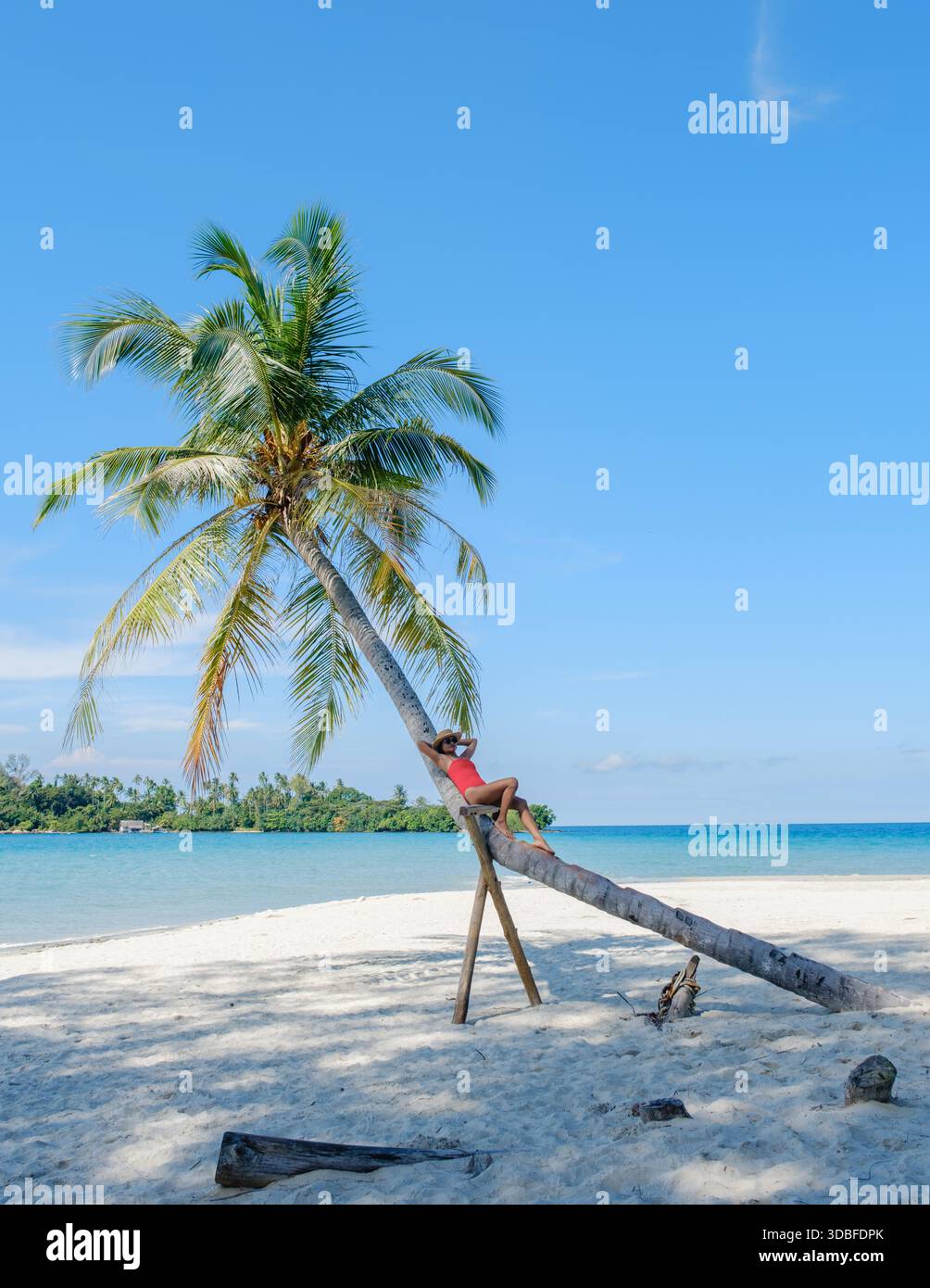 Eine ruhige Szene auf Koh Kood Island zeigt eine asiatische Frau, die auf einer schrägen Palme über weißen Sandstränden liegt. Klarer blauer Himmel und türkisfarbenes Wasser schaffen einen perfekten tropischen Urlaub in Thailand || Model veröffentlicht Stockfoto