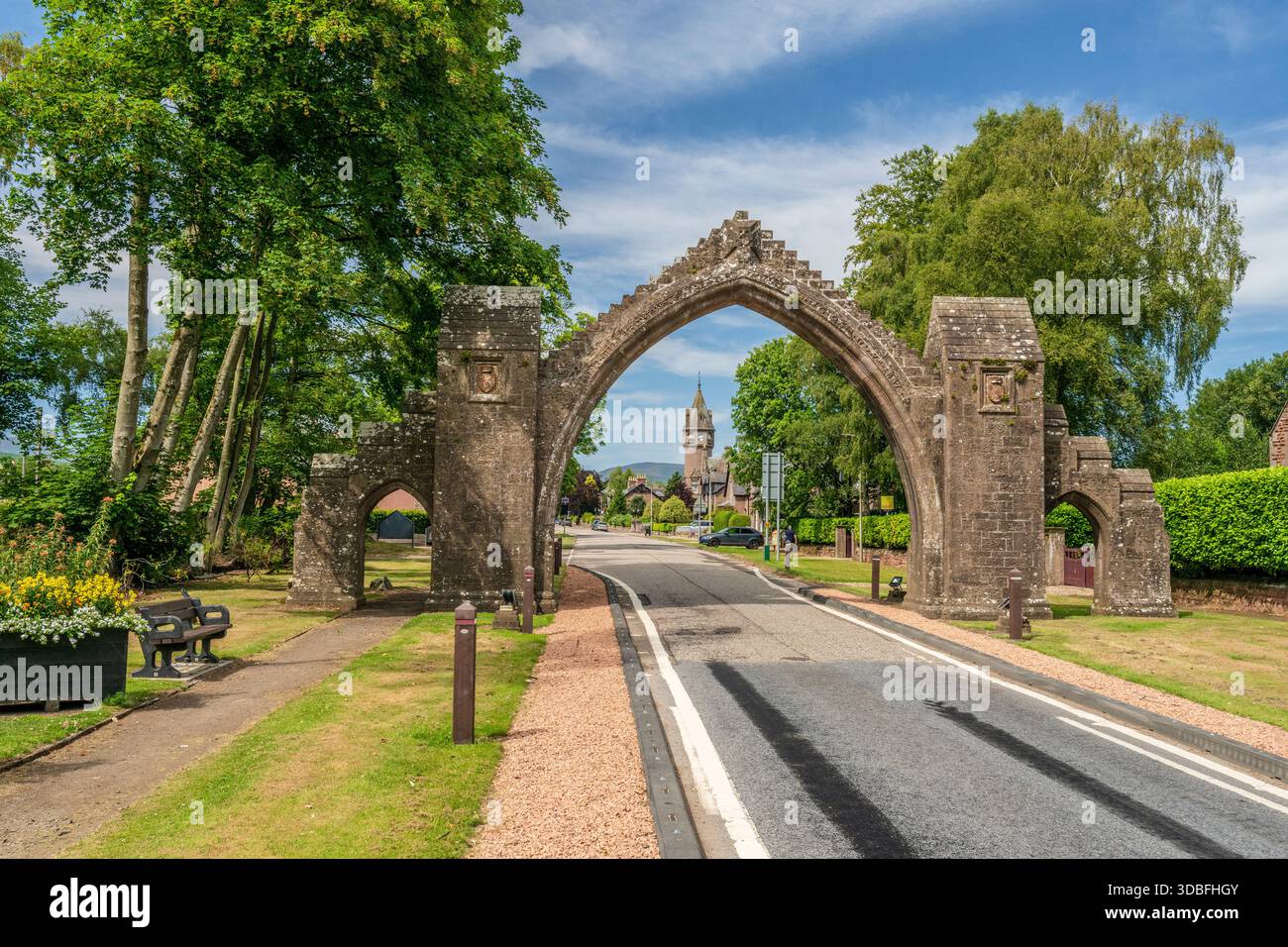 Über einer Landstraße in Edzell, Schottland, erhebt sich ein verwitterter Steinbogen, der von üppigen Bäumen und klarem Himmel eingerahmt wird und an Erbe, zeitlosen Dorfcharme erinnert Stockfoto