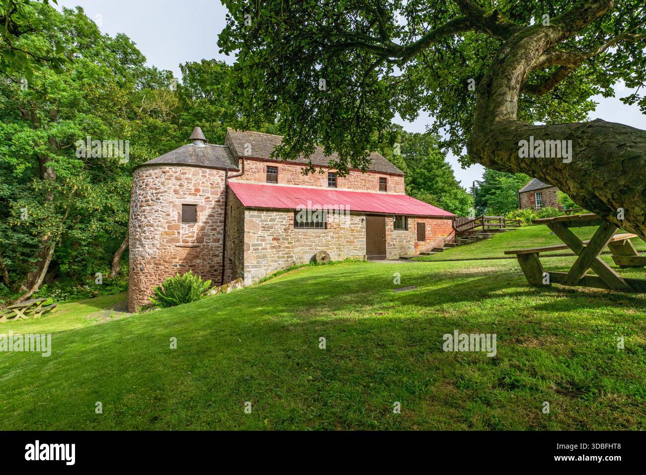 Eine bezaubernde historische Steinmühle mit einem roten Dach befindet sich auf einem grasbewachsenen Rasen, im Schatten eines großen Baumes. Picknicktische laden zu entspannten Besuchen in einem ruhigen ländlichen Raum ein Stockfoto