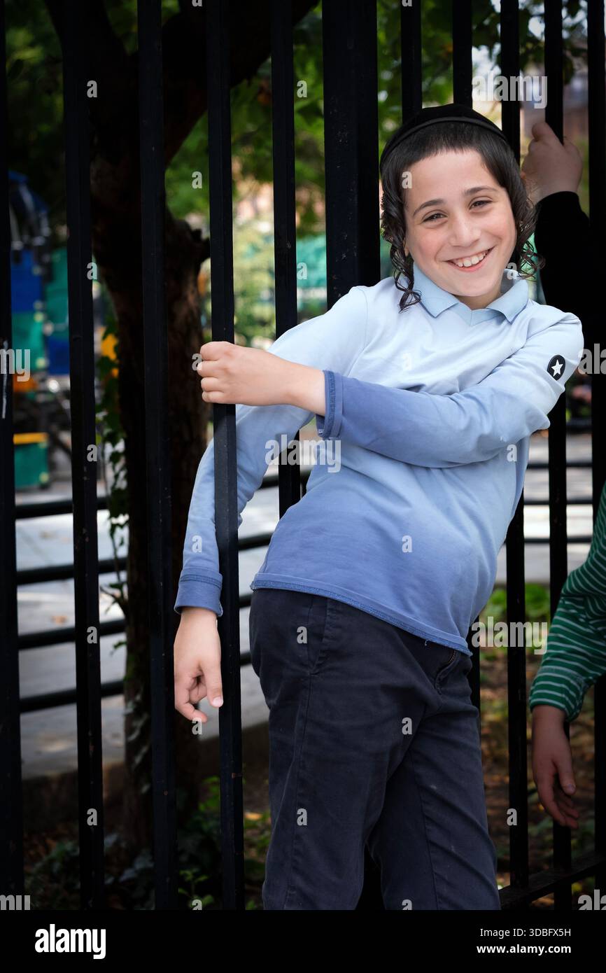 Während der Pause in einer Jeschiwa hängt ein orthodoxer jüdischer Junge in einem Zaun und lächelt. In Brooklyn, New York Stockfoto