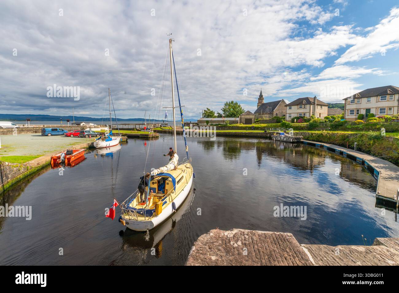 Ein kleines Segelboot ruht in einer von Steinen gesäumten Kanalschleuse am Ardrishaig Basin am Crinan Canal mit Booten, geparkten Autos und einer bezaubernden Skyline des Dorfes Stockfoto