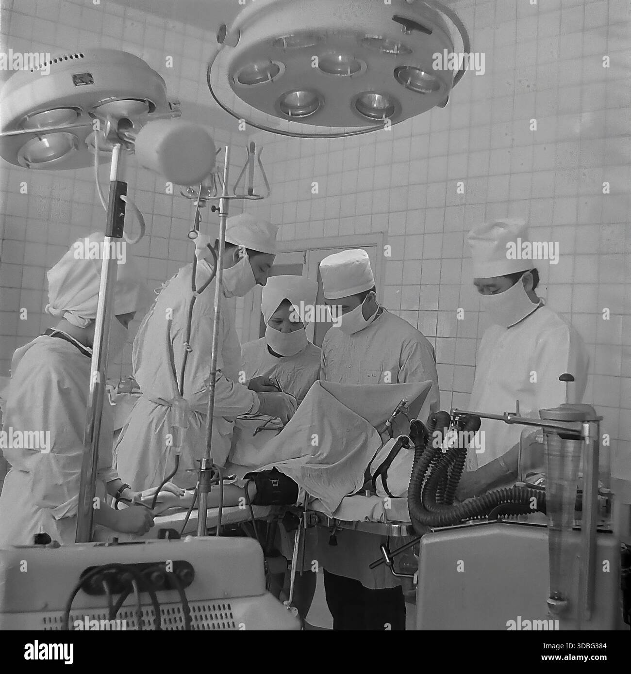 Schwarzweiß-Archivfoto eines Teams von Chirurgen, die eine komplexe Operation in einem Krankenhaus in Donezk (Ukraine, Donbass, 1960er Jahre) durchführen Das medizinische Personal in weißen Kappen und Masken arbeitet mit intensiver Konzentration unter einer großen, schattenlosen Lampe. Im Vordergrund überwacht ein klassisches Anästhesiesystem mit Glasröhren den Patienten. Ein mächtiger Einblick in das fortschrittliche Gesundheitssystem, die berufliche Pflicht und die Rettung von Menschenleben im wissenschaftlichen Zentrum der friedlichen Region lange vor dem Krieg. Stockfoto