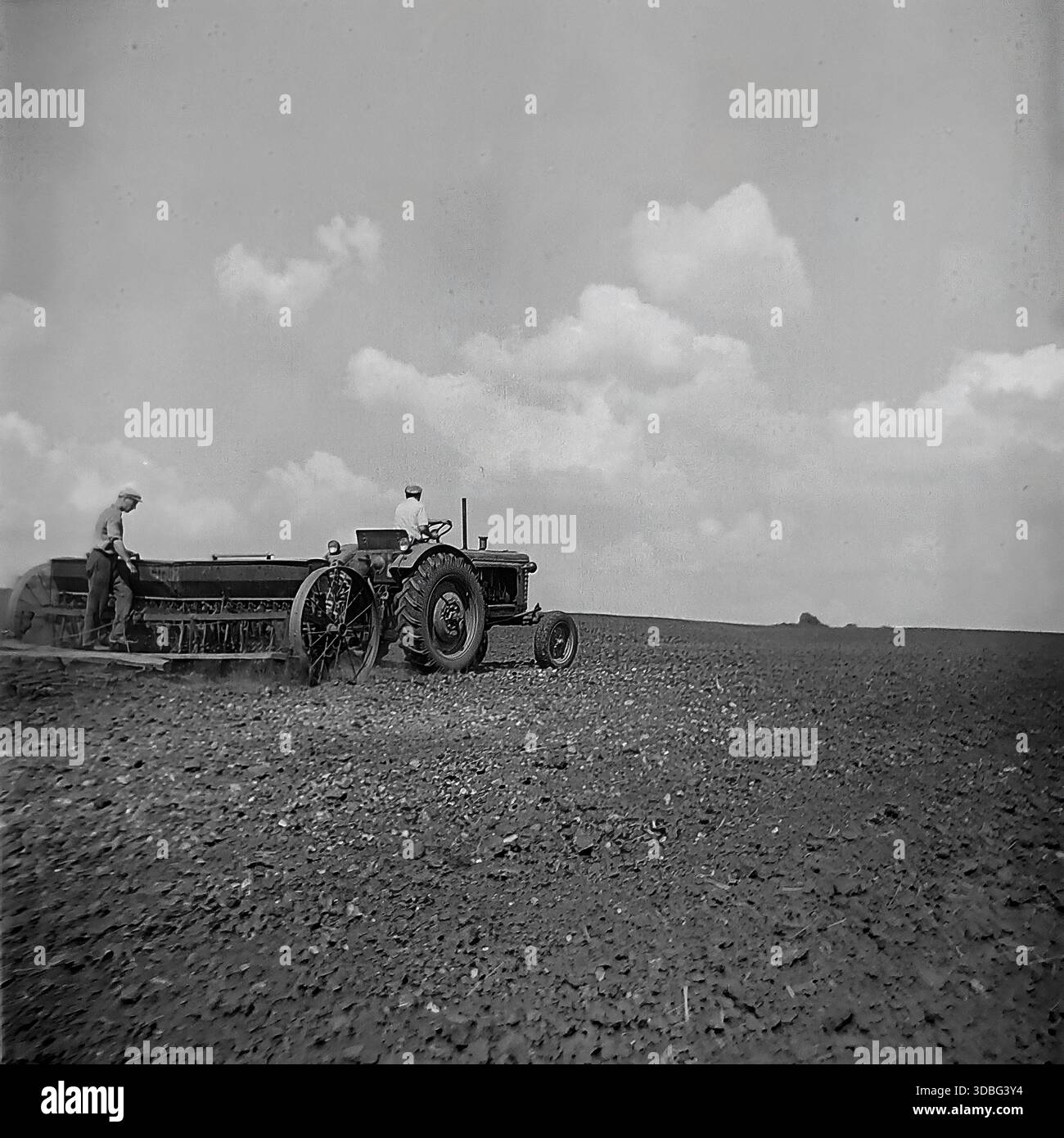 Schwarzweiß-Archivfoto eines Traktors, der auf einem riesigen Feld in der Region Donezk, Ukraine, Donbass, 1960er Jahre arbeitet Zwei männliche Arbeiter bewirtschaften die schweren Maschinen, säen Samen oder begraben den fruchtbaren schwarzen Boden während des Frühlingsfeldzugs. Der Traktor zieht ein breites landwirtschaftliches Arbeitsgerät, das unter bewölktem Himmel Staub aufwirbelt. Eine mächtige Szene sowjetischer mechanisierter Landwirtschaft, kollektiver Arbeit und der endlosen Horizonte der friedlichen Agrarlandschaft vor dem Krieg. Stockfoto
