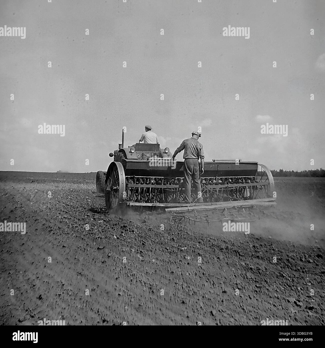 Schwarzweiß-Archivfoto eines Traktors, der auf einem riesigen Feld in der Region Donezk, Ukraine, Donbass, 1960er Jahre arbeitet Zwei männliche Arbeiter bewirtschaften die schweren Maschinen, säen Samen oder begraben den fruchtbaren schwarzen Boden während des Frühlingsfeldzugs. Der Traktor zieht ein breites landwirtschaftliches Arbeitsgerät, das unter bewölktem Himmel Staub aufwirbelt. Eine mächtige Szene sowjetischer mechanisierter Landwirtschaft, kollektiver Arbeit und der endlosen Horizonte der friedlichen Agrarlandschaft vor dem Krieg. Stockfoto