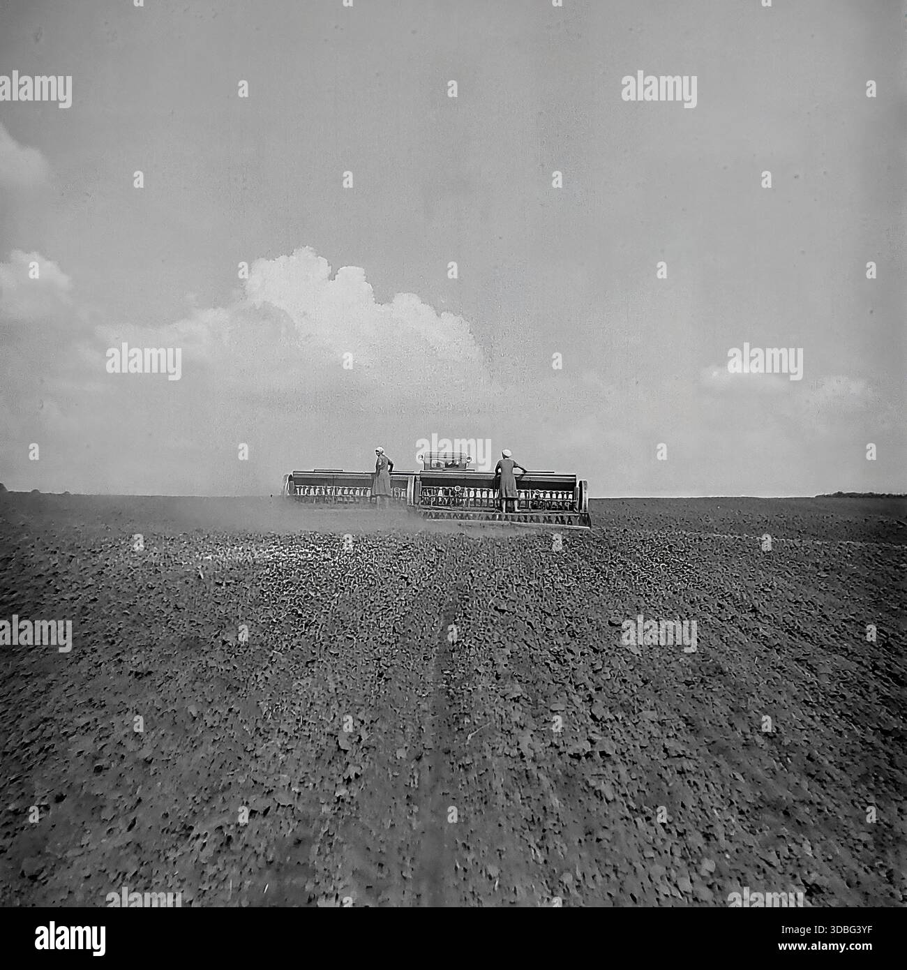 Schwarzweiß-Archivfoto eines Traktors, der auf einem riesigen Feld in der Region Donezk, Ukraine, Donbass, 1960er Jahre arbeitet Zwei männliche Arbeiter bewirtschaften die schweren Maschinen, säen Samen oder begraben den fruchtbaren schwarzen Boden während des Frühlingsfeldzugs. Der Traktor zieht ein breites landwirtschaftliches Arbeitsgerät, das unter bewölktem Himmel Staub aufwirbelt. Eine mächtige Szene sowjetischer mechanisierter Landwirtschaft, kollektiver Arbeit und der endlosen Horizonte der friedlichen Agrarlandschaft vor dem Krieg. Stockfoto
