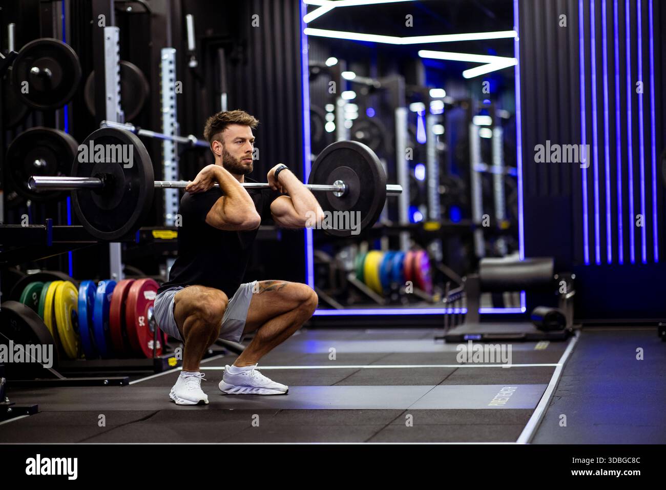 In einem modernen Fitnessstudio mit bunten Gewichten führt ein Mann eine Langhantelhocke aus. Er konzentriert sich intensiv und zeigt Stärke und richtige Form unter bri Stockfoto
