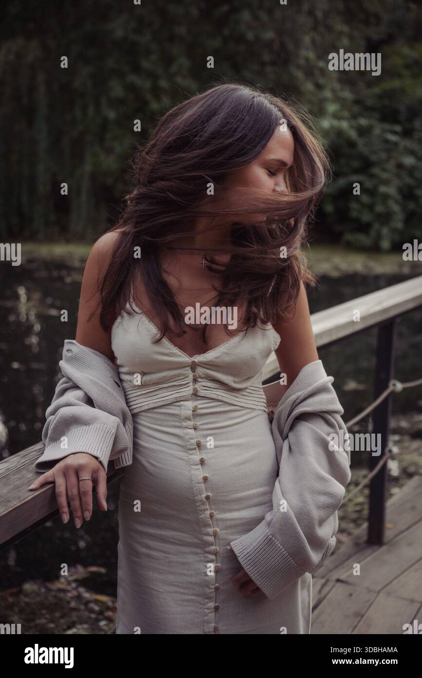 Junge Frau in Kleid und Strickjacke steht auf einer Holzbrücke am Teich, Wind bewegt ihre Haare, sanftes Tageslicht, gedämpfte Grüntöne, spontane Stimmung Stockfoto