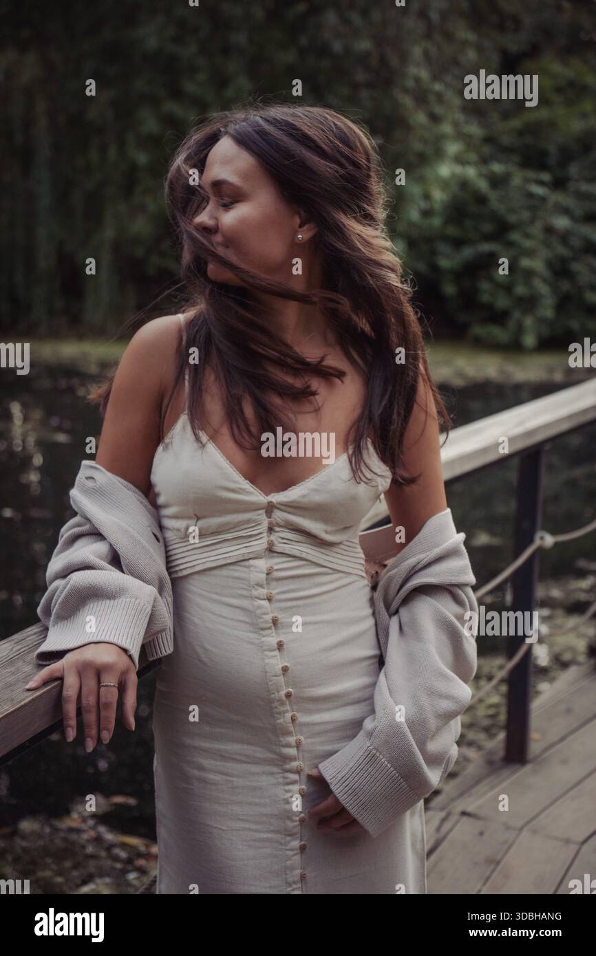 Junge Frau in Kleid und Strickjacke steht auf einer Holzbrücke am Teich, Wind bewegt ihre Haare, sanftes Tageslicht, gedämpfte Grüntöne, spontane Stimmung Stockfoto