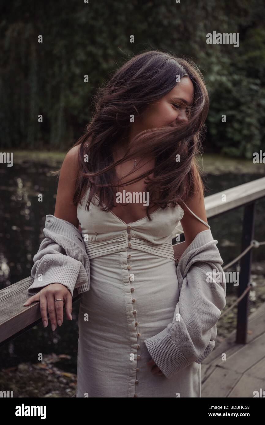Junge Frau in Kleid und Strickjacke steht auf einer Holzbrücke am Teich, Wind bewegt ihre Haare, sanftes Tageslicht, gedämpfte Grüntöne, spontane Stimmung Stockfoto
