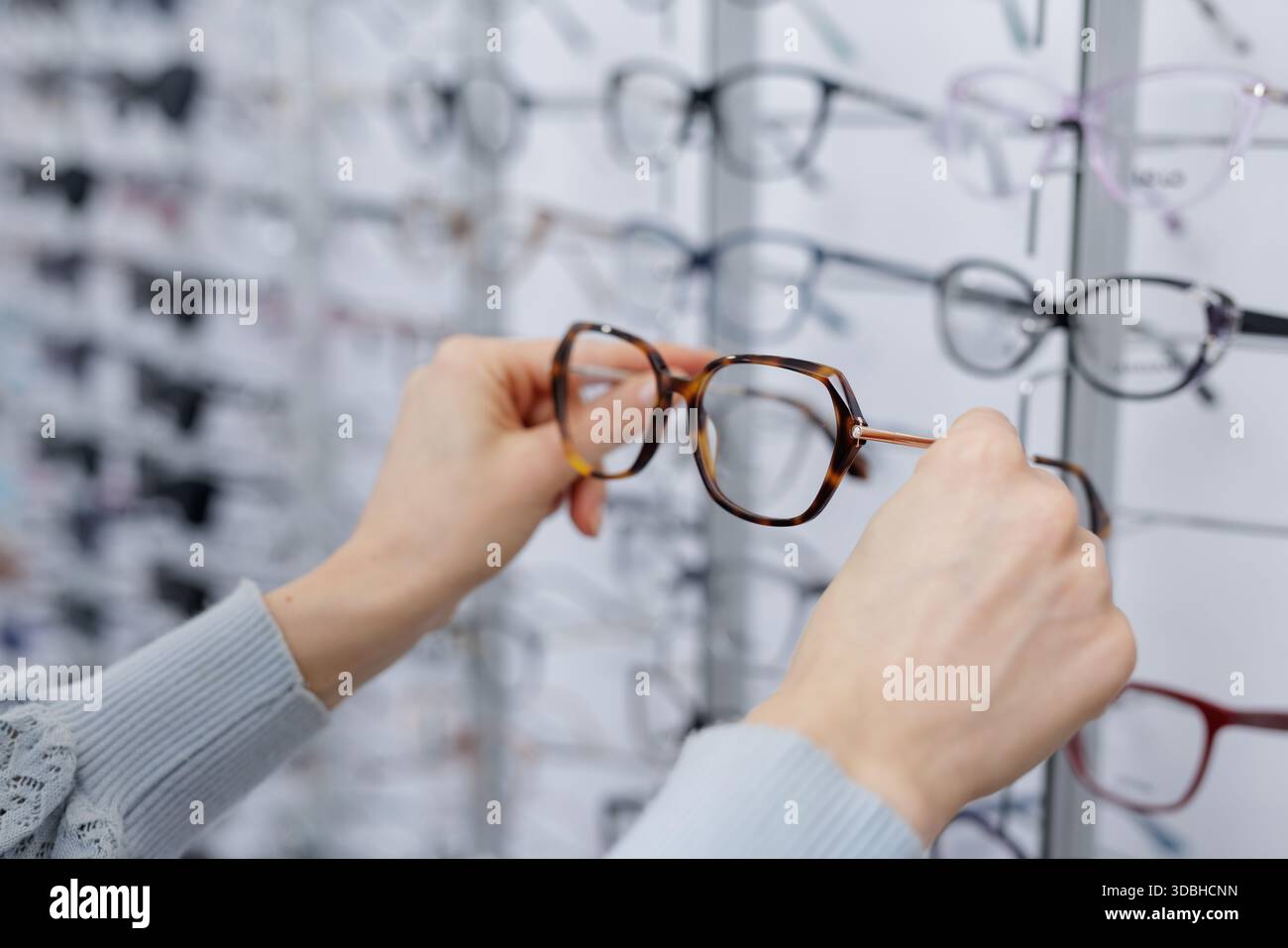 Hände halten Schildpatt-Brillen im Optikerausstellungsraum und wählen Brillen aus der Displaywand Stockfoto