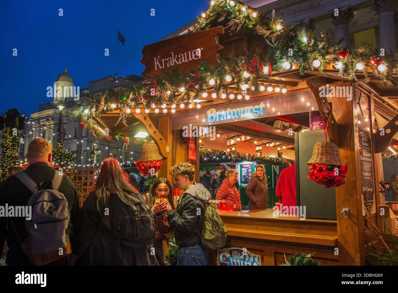 Ein junges Paar genießt einen Hotdog von einem Stand auf der Weihnachtsmesse am Trafalgar Square, London. Weihnachtsstände. Essen. Stockfoto
