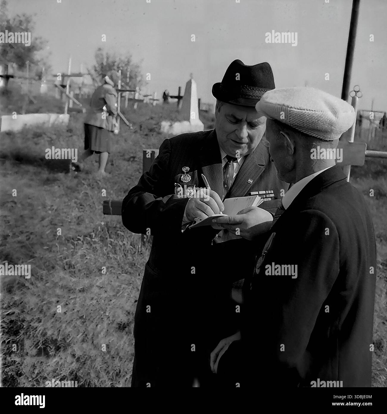 September 1978. Dorf Dolina, Bezirk Sloviansk, Region Donezk, ukrainische SSR, UdSSR. Zwei ältere Männer, wahrscheinlich Veteranen des Zweiten Weltkriegs, unterhalten sich auf einem örtlichen Friedhof während des 35. Jahrestages der Befreiung des Dorfes von der Nazi-Besatzung. Ein Mann, der einen Fedorahut und viele Dienstmedaillen auf seiner dunklen Anzugsjacke trägt, schreibt Kontaktdaten in ein kleines Notizbuch, während der andere ansieht. Im Hintergrund spaziert eine Frau zwischen den hölzernen Grabkreuzen. Authentisches Archivbild, das die Atmosphäre der Erinnerung, der Bruderschaft und des friedlichen Lebens im sowjetischen Donbass erfasst. Stockfoto