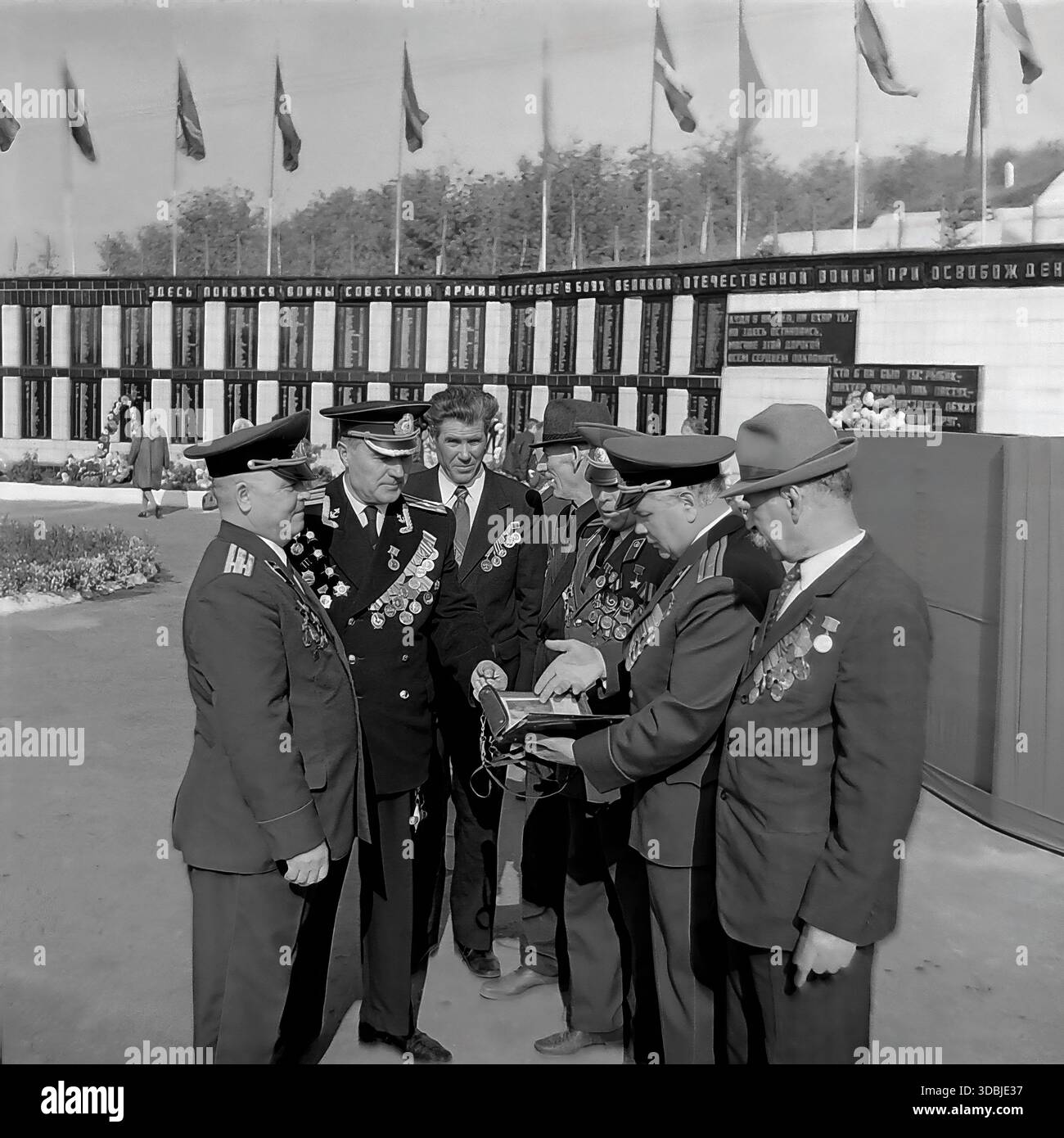 September 1978. Dorf Dolina, Bezirk Sloviansk, Region Donezk, ukrainische SSR, UdSSR. Während des 35. Jahrestages der Befreiung des Dorfes von der Nazi-Besatzung versammeln sich in der Nähe der Gedenkmauer eine Gruppe dekorierter Veteranen des Zweiten Weltkriegs, darunter Militäroffiziere und Zivilisten. Die Kameraden untersuchen ein Erinnerungsalbum, während sie unter Fahnen stehen. Authentisches Archivbild, das die Atmosphäre der Brüderlichkeit, des Patriotismus und des friedlichen Lebens im sowjetischen Donbass während der Breschnew-Ära feststellt. Stockfoto