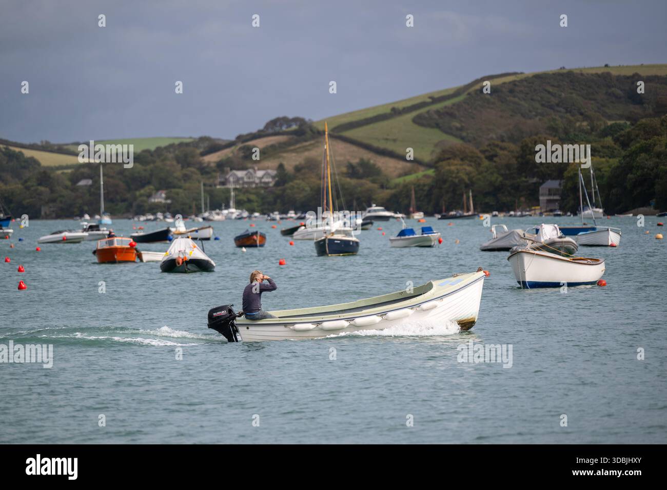Weißer Mann, der seine Augen beschattet, während er ein Schlauchboot über den Hafen von Salcombe fährt, mit einem Außenbordmotor, der im Herbst eine kleine Spur erzeugt Stockfoto