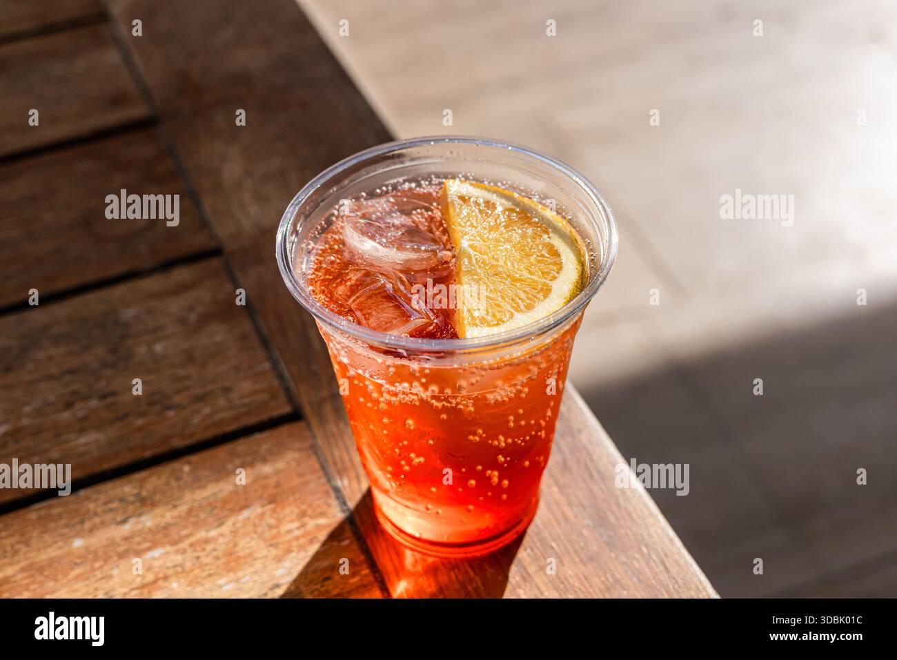 Ein erfrischendes Getränk in einem klaren Plastikbecher, gefüllt mit prickelndem orangenem Limonade. Der Becher wird auf einen Holztisch mit verschwommenem Hintergrund gestellt. Stockfoto
