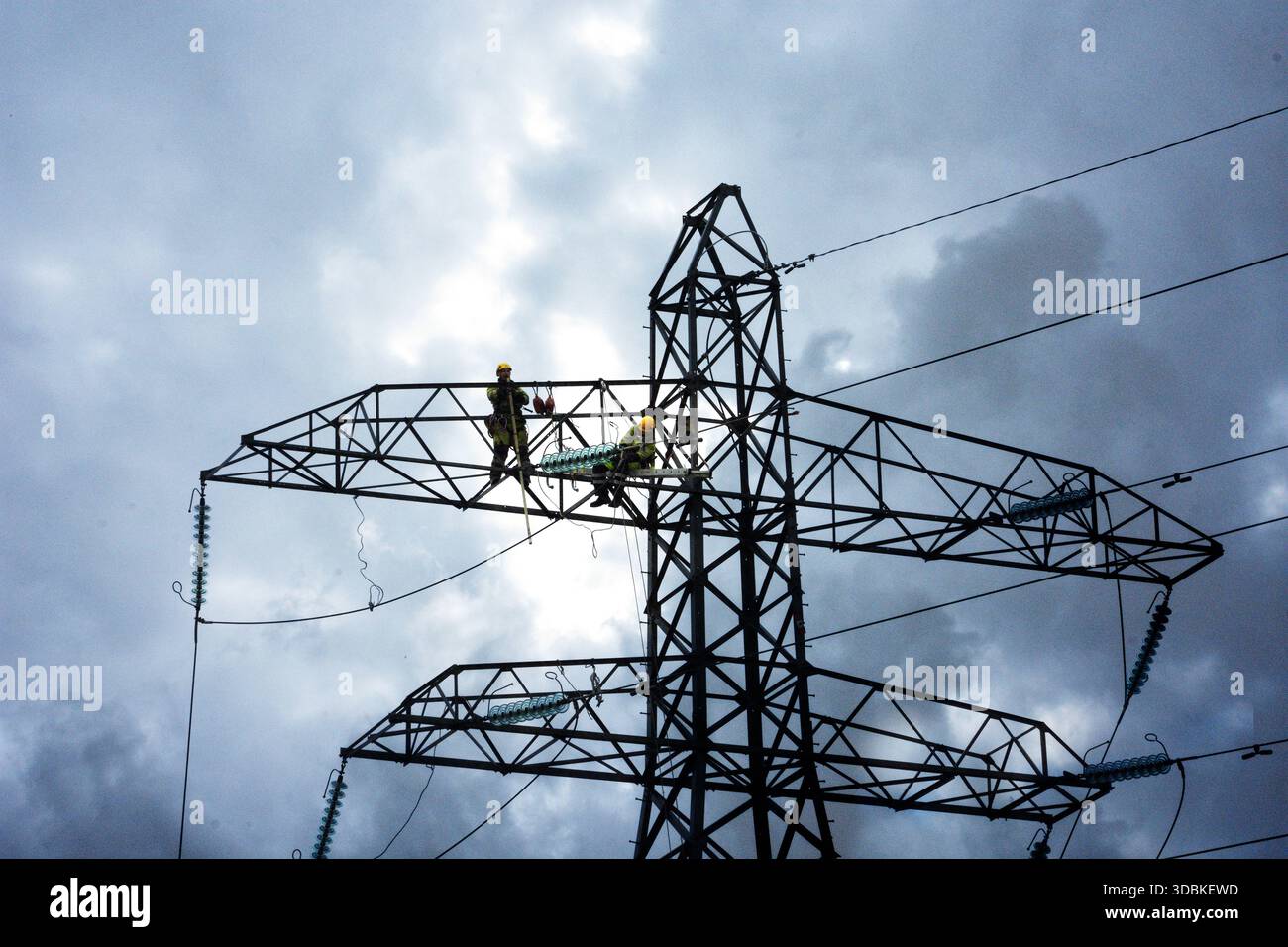 Zwei Versorgungsarbeiter führen Wartungsarbeiten auf hohem Niveau an einem Stromübertragungspylon durch und betonen die Skalenkomplexität Stockfoto