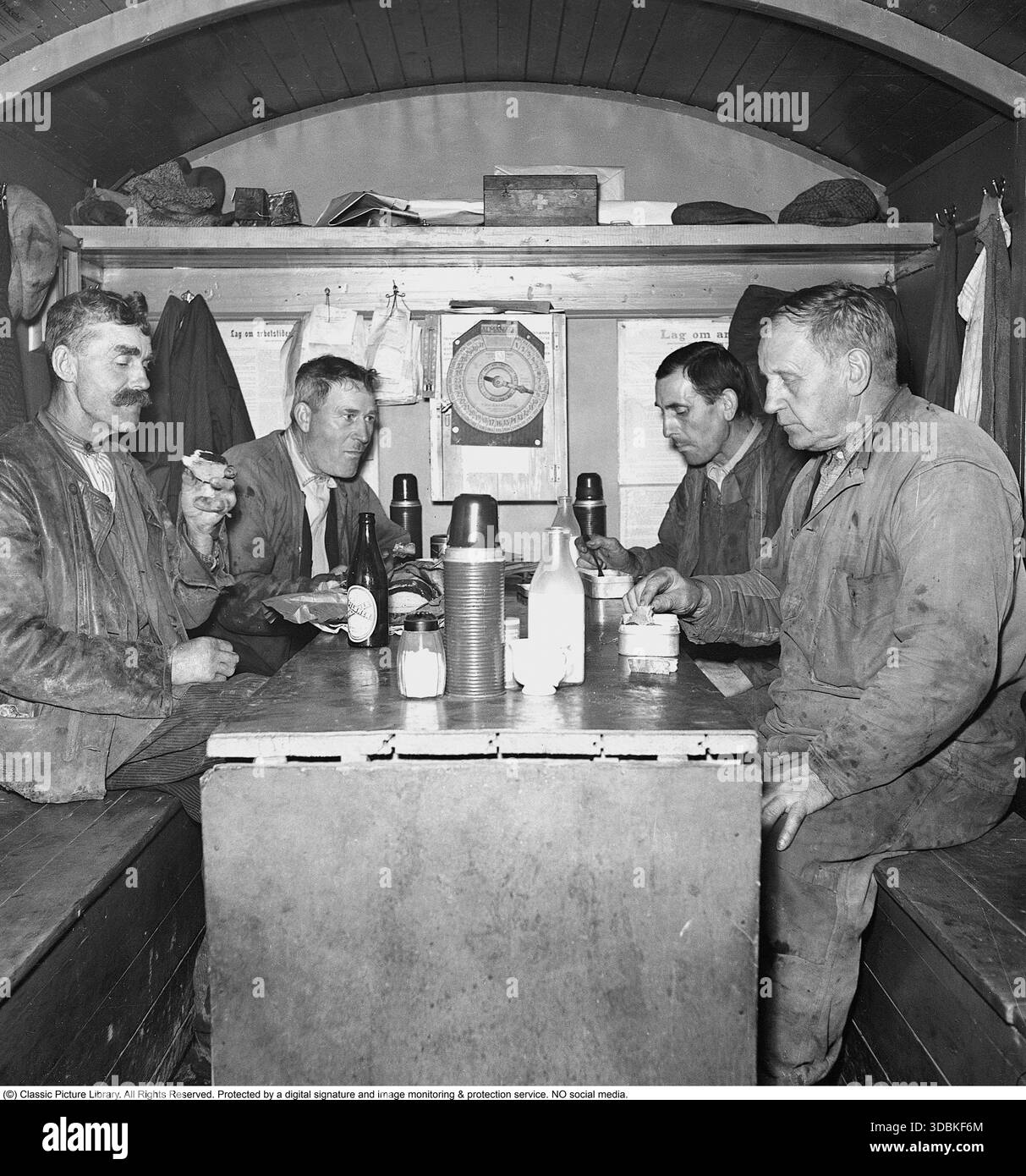 Mittagspause für die Jungs (1942) es ist Mittagspause im Jahr 1942, und vier Jungs von Stockholms Straßenpflegepersonal machen eine wohlverdiente Pause in ihrer mobilen Hütte – einer tragbaren Kabine, die auch als Umkleidekabine und Kantine dient. Immer noch in ihrer schmuddeligen Arbeitskleidung, mit schwarzen Händen vom Job, verstauen sie sich hausgemachte Mahlzeiten aus klassischen Metalldosen, die mit einer Flasche Milch oder einem Pilsner-Bier abgespült werden. In den frühen 1940er Jahren, vor allem während der Kriegsrationierung, verließen sich Handarbeiter in ganz Europa (einschließlich des neutralen Schwedens) auf stabile Metalldosen, die mit einfachen, deftigen Speisen gefüllt waren Stockfoto