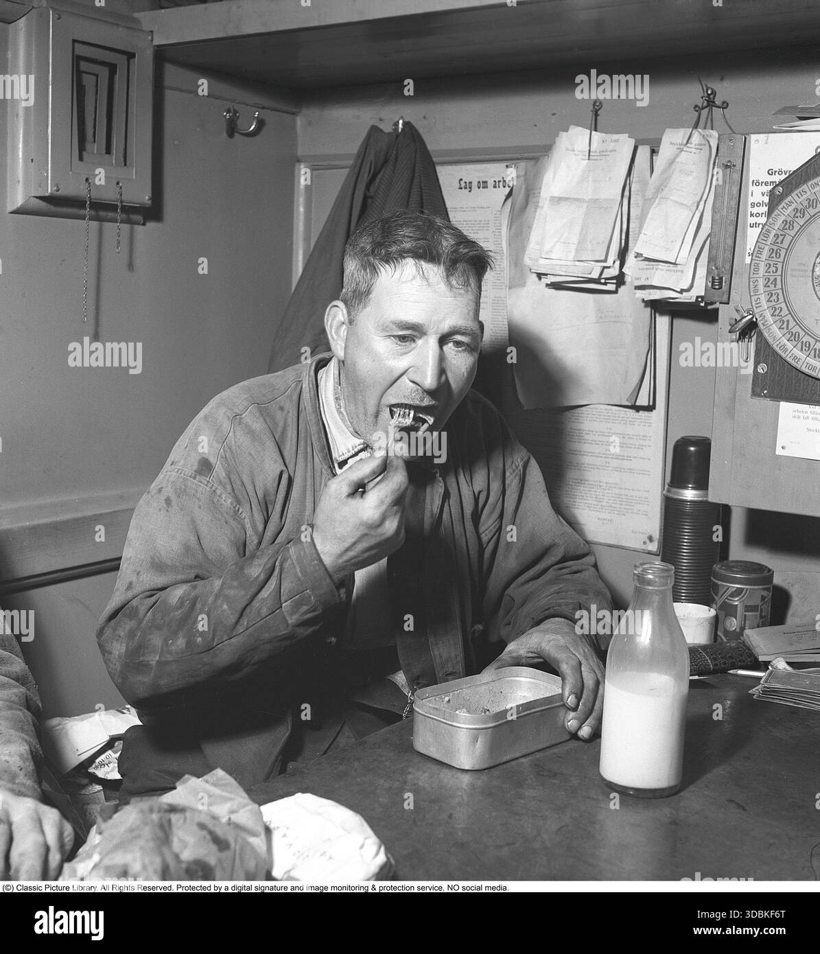 Mittagspause für die Jungs (1942) es ist Mittagspause im Jahr 1942, und Jungs von Stockholms Straßenpflegepersonal machen eine wohlverdiente Pause in ihrer mobilen Hütte – einer tragbaren Kabine, die auch als Umkleidekabine und Kantine dient. Immer noch in ihrer schmuddeligen Arbeitskleidung, mit schwarzen Händen vom Job, verstauen sie sich hausgemachte Mahlzeiten aus klassischen Metalldosen, die mit einer Flasche Milch oder einem Pilsner-Bier abgespült werden. In den frühen 1940er Jahren, besonders während der Kriegsrationierung, verließen sich Handarbeiter in ganz Europa (einschließlich des neutralen Schweden) auf stabile Metalldosen, die mit einfachen, deftigen Speisen gefüllt waren Stockfoto