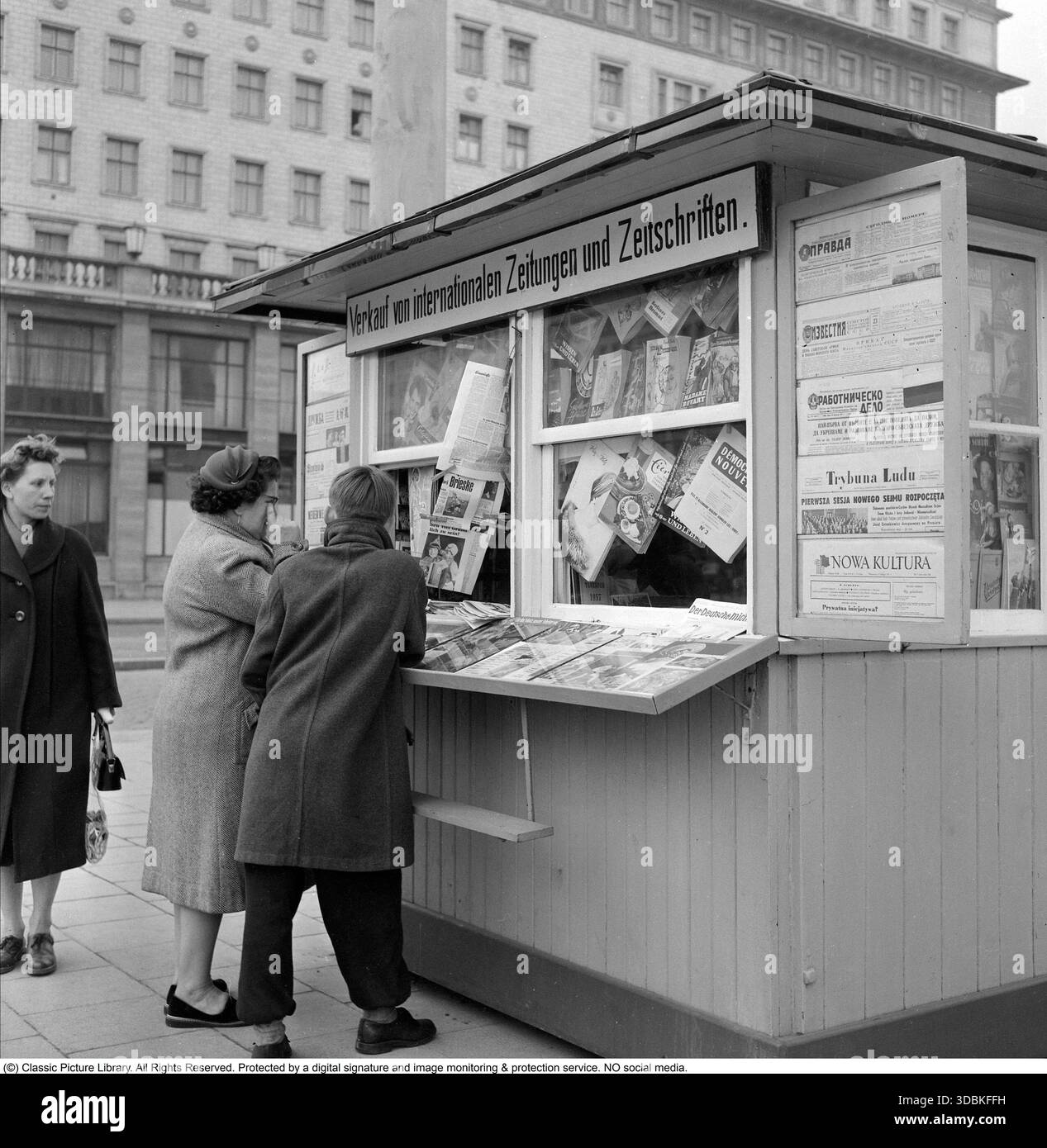 International Newspapers Kiosk, Ost-Berlin (1957) dieses Foto aus dem Jahr 1957 zeigt einen Straßenkiosk in Ost-Berlin, der stolz den Verkauf von internationalen Zeitungen und Zeitschriften anpreist – ein seltenes Fenster zur weiteren Welt in der streng kontrollierten DDR. In den 1950er Jahren waren westliche Publikationen stark eingeschränkt: Gewöhnliche Bürger hatten wenig bis gar keinen Zugang, die ausländische Presse beschränkte sich auf Parteifunktionäre, Institutionen und anerkannte Eliten, um eine „ideologische Kontamination“ zu verhindern. Diese speziellen Kioske wurden sorgfältig ausgewählt Stockfoto