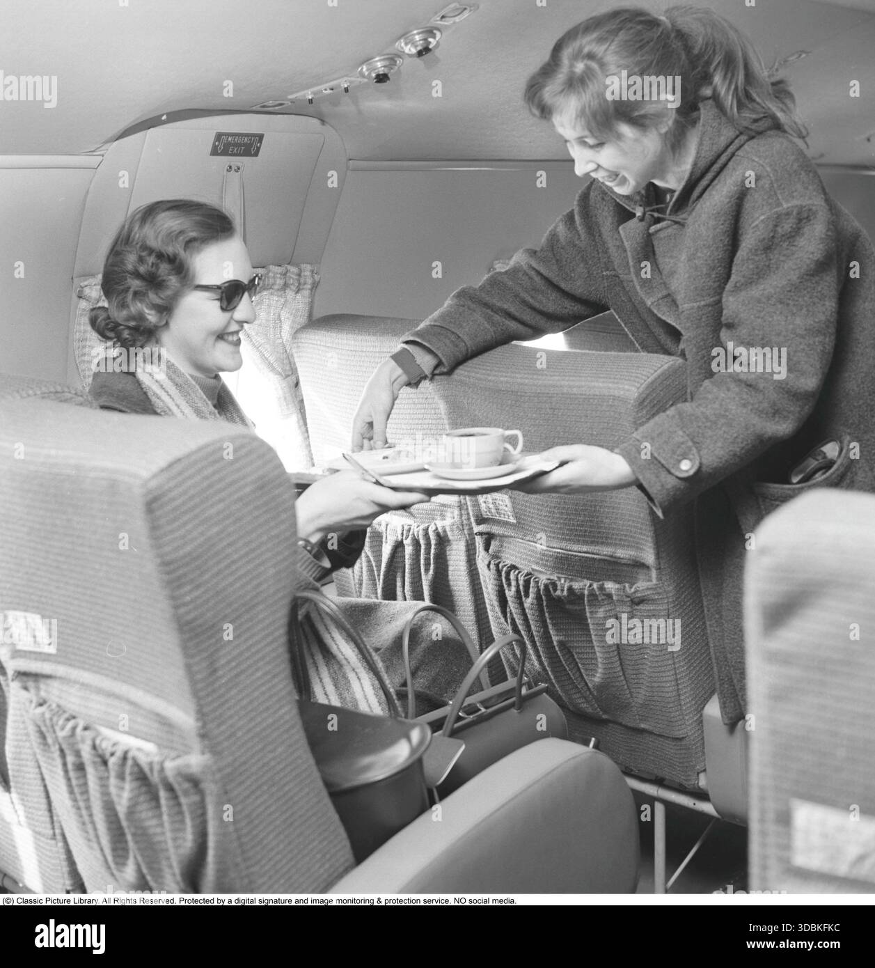 Flugreisen in den 1950er Jahren Das weibliche Mitglied der Crew serviert den Passagieren Kaffee und Sandwiches. 1957. Das weibliche Besatzungsmitglied trägt einen Duffle-Mantel, einen warmen Kapuzenmantel, der traditionell aus dickem Wollgewebe hergestellt wird und mit markanten Knebelverschlüssen aus Holz oder Horn und großen aufgesetzten Taschen ausgestattet ist. Aus belgischem Seetuch stammend und im späten 19. Jahrhundert von der britischen Royal Navy populär gemacht, wurde es in den 1950er–1960er Jahren wegen seiner praktischen und lässigen Art zu einem zivilen Modeartikel. Stockfoto