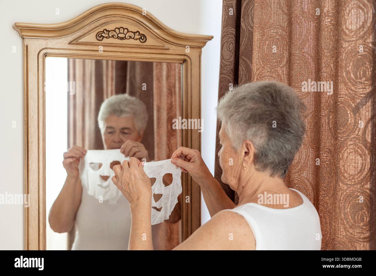 Ältere Frau, die zu Hause Hautpflegemaske vor dem Spiegel aufträgt – Schönheit, Selbstpflege und Anti-Aging-Routine für reife Haut und Wellness-Konzept Stockfoto