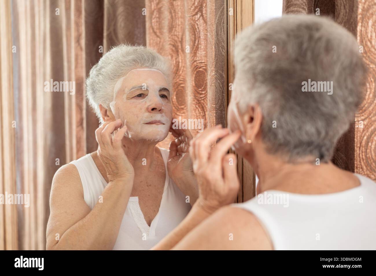 Ältere Frau, die zu Hause Hautpflegemaske vor dem Spiegel aufträgt – Schönheit, Selbstpflege und Anti-Aging-Routine für reife Haut und Wellness-Konzept Stockfoto