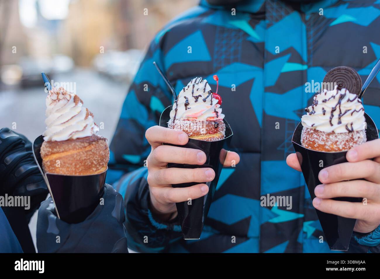 Drei Schornsteinkuchen, gehalten von einer Person in einer Winterjacke. Jeder Kuchen variiert: Einer mit weißer Sahne und Streuseln, ein anderer mit Sahne, Schokoladenbelag, Stockfoto