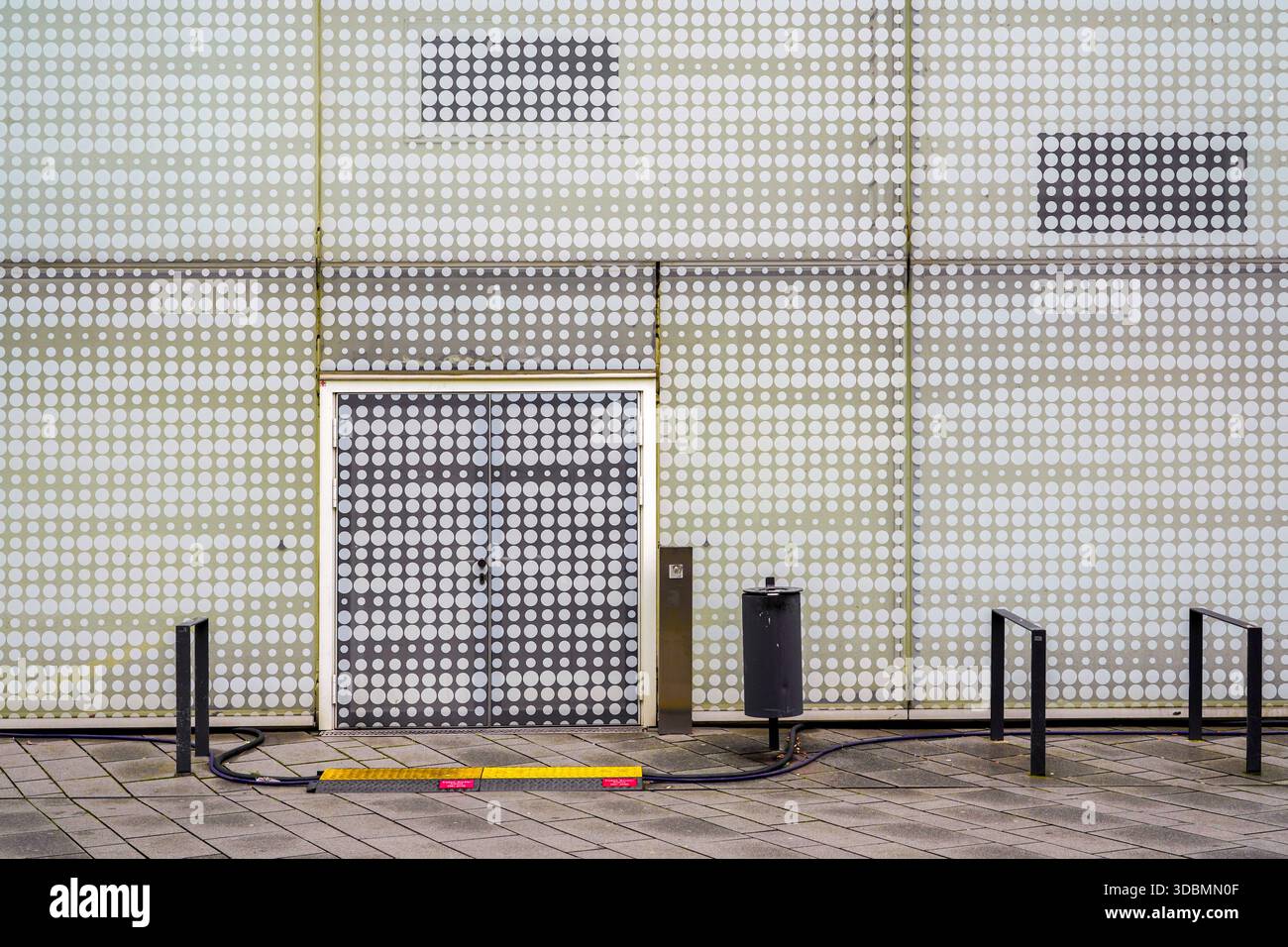 Rhythmische Wiederholung: Polka-Doted Pattern. Ein Stück Moderne im Forum Confluentes in Koblenz, wo Kreise und Quadrate in monochromatischen Tönen eine visuelle Symphonie schaffen, die von Benthem Crouwel Architects orchestriert wurde. Das gepunktete Muster der Fassade wird durch eine kontrastierende Tür segmentiert. Der umliegende Poller, Fahrradständer, Aschenbecher und der Bürgersteig ergänzen die moderne Atmosphäre des Gebäudes. Stockfoto