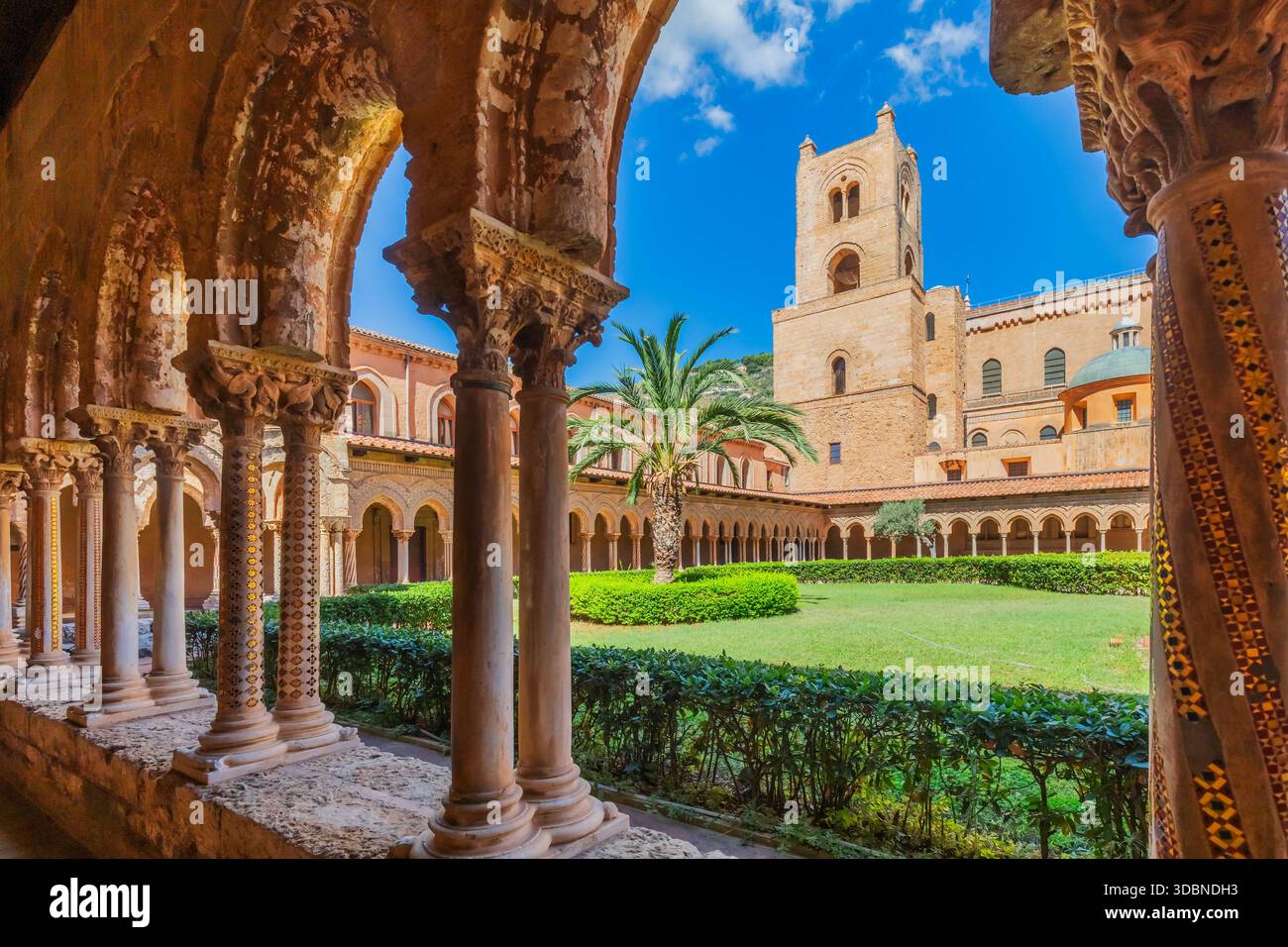 Blick auf den Benediktinerkloster von Monreale, mit kunstvollen Steinbögen, geschnitzten Kapitellen, Säulen, Palmen und dem mittelalterlichen Glockenturm der Kathedrale von Monreale, einem Meisterwerk normannischer Architektur. Metropolstadt Palermo, Sizilien, Italien Stockfoto