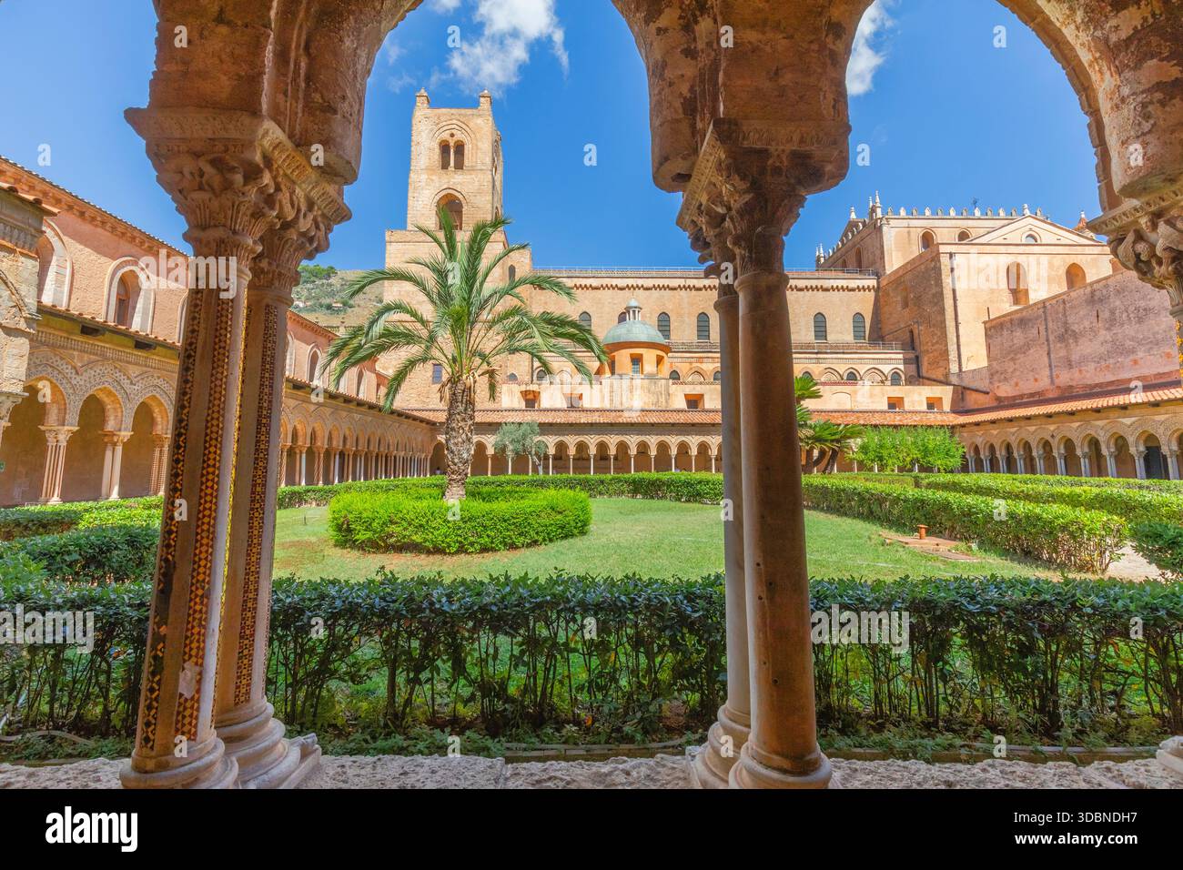 Blick auf den Benediktinerkloster von Monreale, mit kunstvollen Steinbögen, geschnitzten Kapitellen, Säulen, Palmen und dem mittelalterlichen Glockenturm der Kathedrale von Monreale, einem Meisterwerk normannischer Architektur. Metropolstadt Palermo, Sizilien, Italien Stockfoto