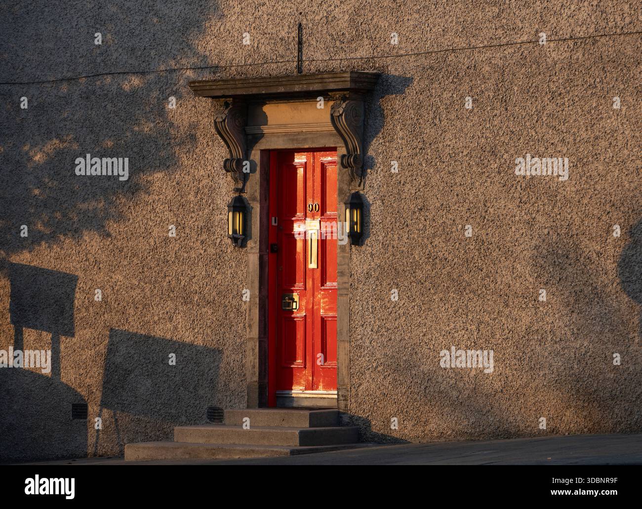 Rote Tür mit kunstvollem Steinrahmen, Laternen und warmen Schatten an einer strukturierten Wand. Stockfoto