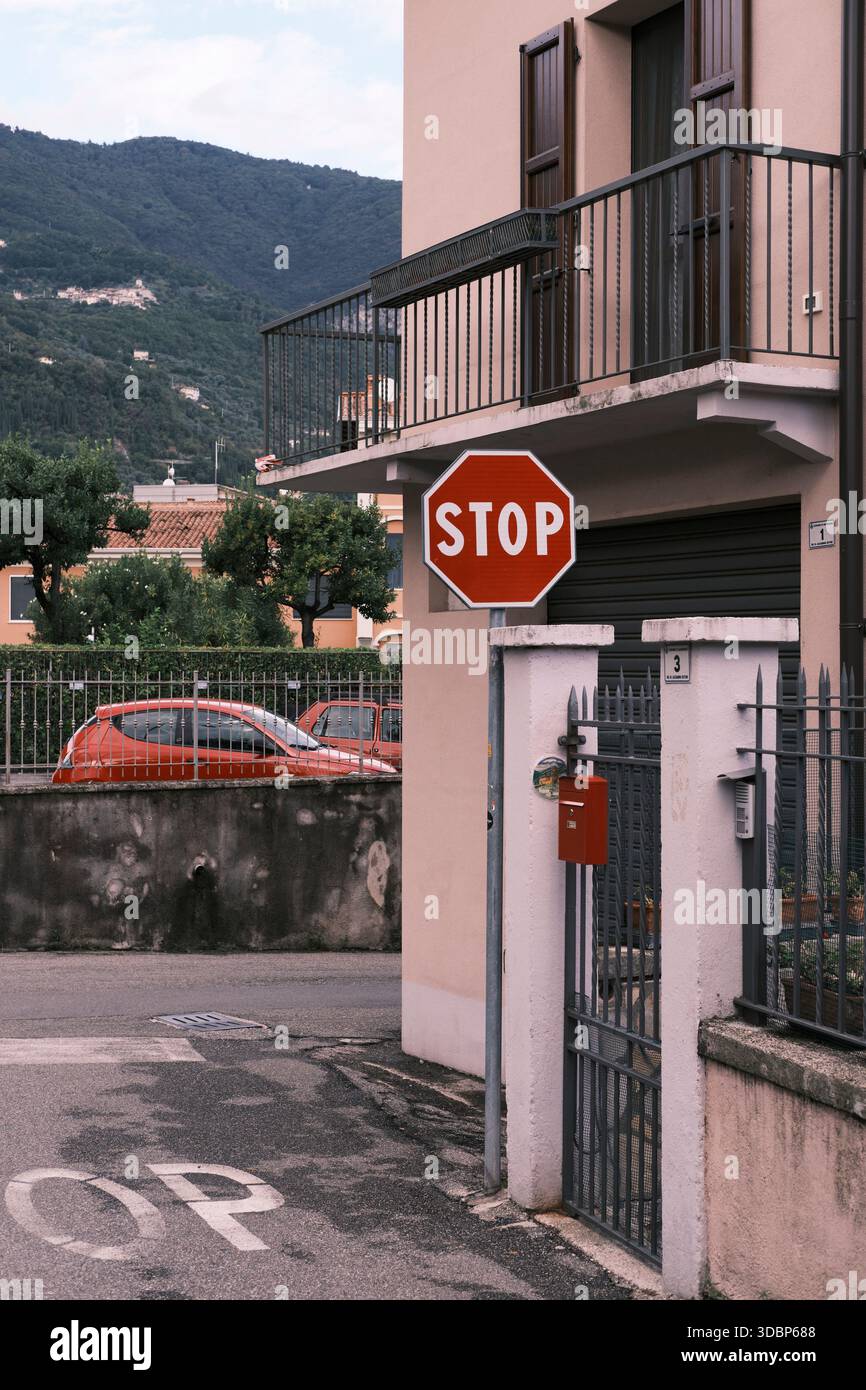 Seitenstraße in Bogliaco am Gardasee. Rotes Stoppschild neben einem roten Briefkasten. Zwei rote Autos parkten im Hintergrund. Stockfoto