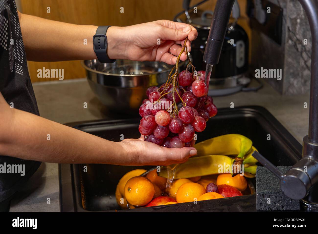 Frische Trauben und Obst im Spülbecken waschen. Person, die rote Trauben unter fließendem Wasser in einer Küchenspüle mit Bananen, Orangen und spült Stockfoto