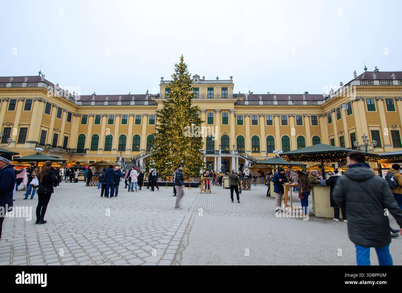 Die Leute spazieren und shoppen auf einem festlichen Weihnachtsmarkt mit Lichtern und Dekorationen vor einem großen historischen Gebäude mit Bogenfenstern Stockfoto