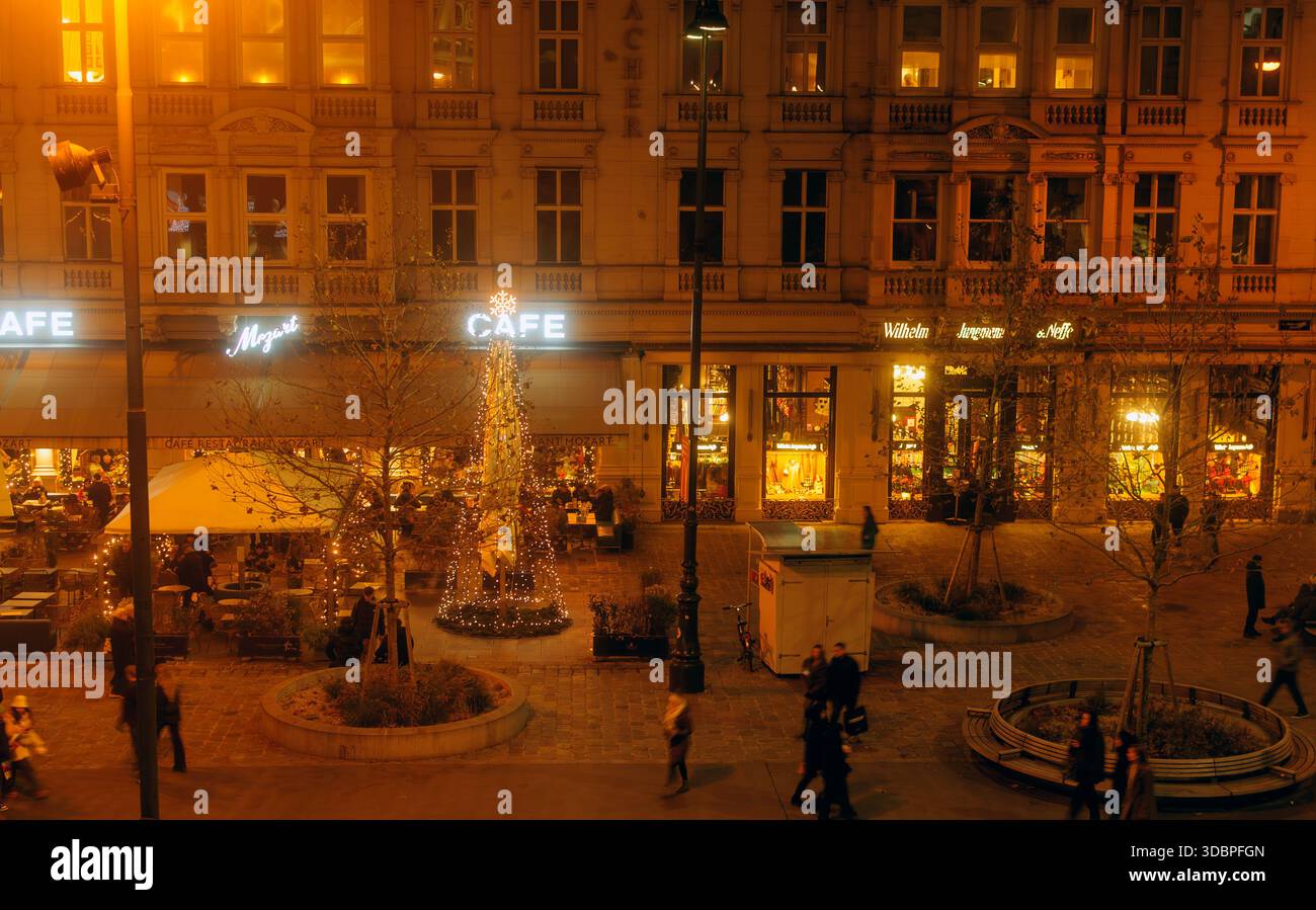 Abendliche Straßenszene mit Leuten, die an beleuchteten Cafés und einem geschmückten Weihnachtsbaum in festlicher urbaner Umgebung vorbeilaufen Stockfoto