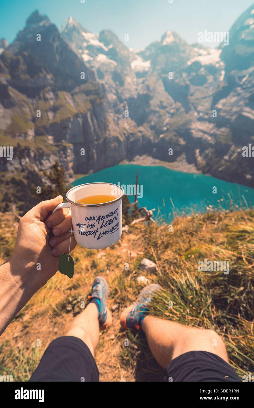 Jemand hält eine Tasse Tee in der Hand, während unten der malerische Oeschinensee in der Mitte der Schweizer Berge zu sehen ist. Die Teetasse ist mit dem Zitat „ohne Abenteuer, das Leben wäre tödlich langweilig“ bedruckt. Stockfoto