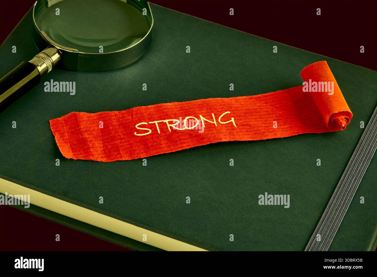 Eine Lupe sitzt neben einer orangefarbenen Schriftrolle, die das Wort STARK trägt. Die Rolle liegt auf einem geschlossenen, dunkelgrünen Journal mit elastischem Verschluss. Stockfoto